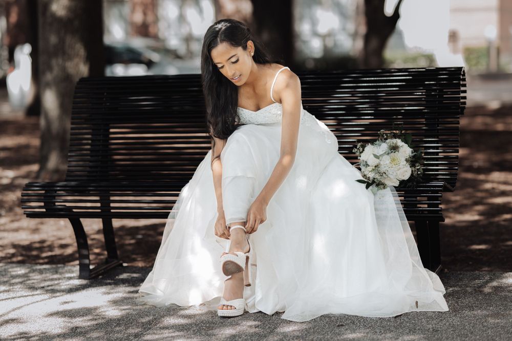 A bride in a flowing white gown adjusts her dress in a shaded outdoor setting.