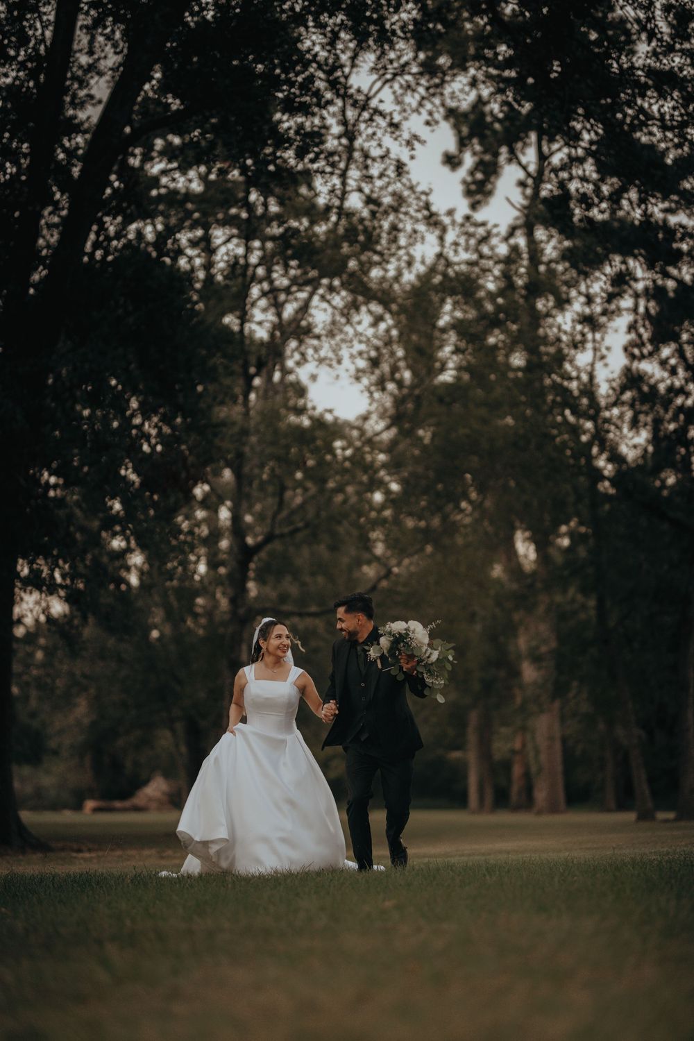 Wedding couple walking together on grassy field with trees silhouetted at dusk.