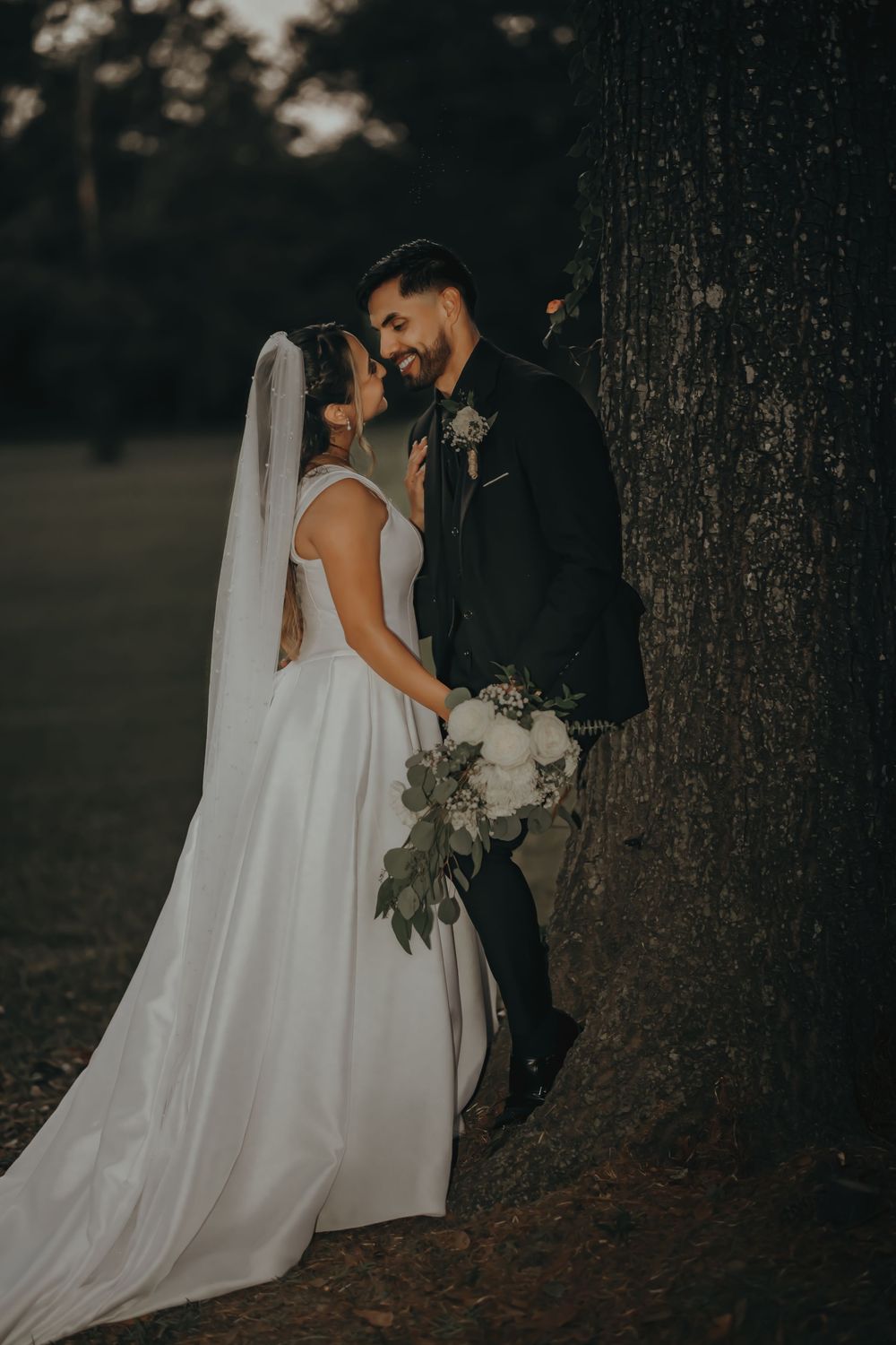 Romantic wedding couple embraces in a moody forest setting with flowing white dress and dark suit.