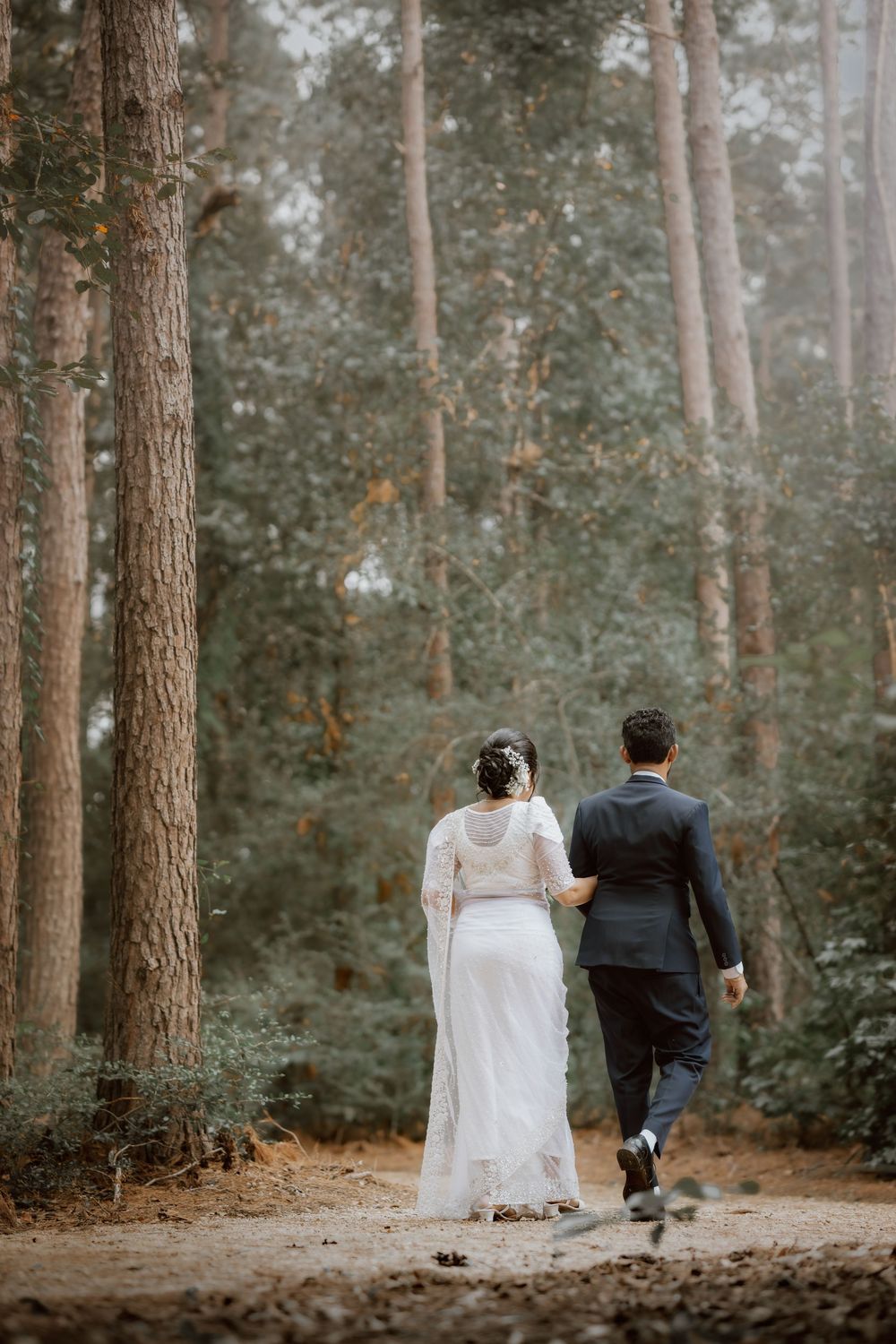 A bride and groom walk together through a misty pine forest path during their wedding day.