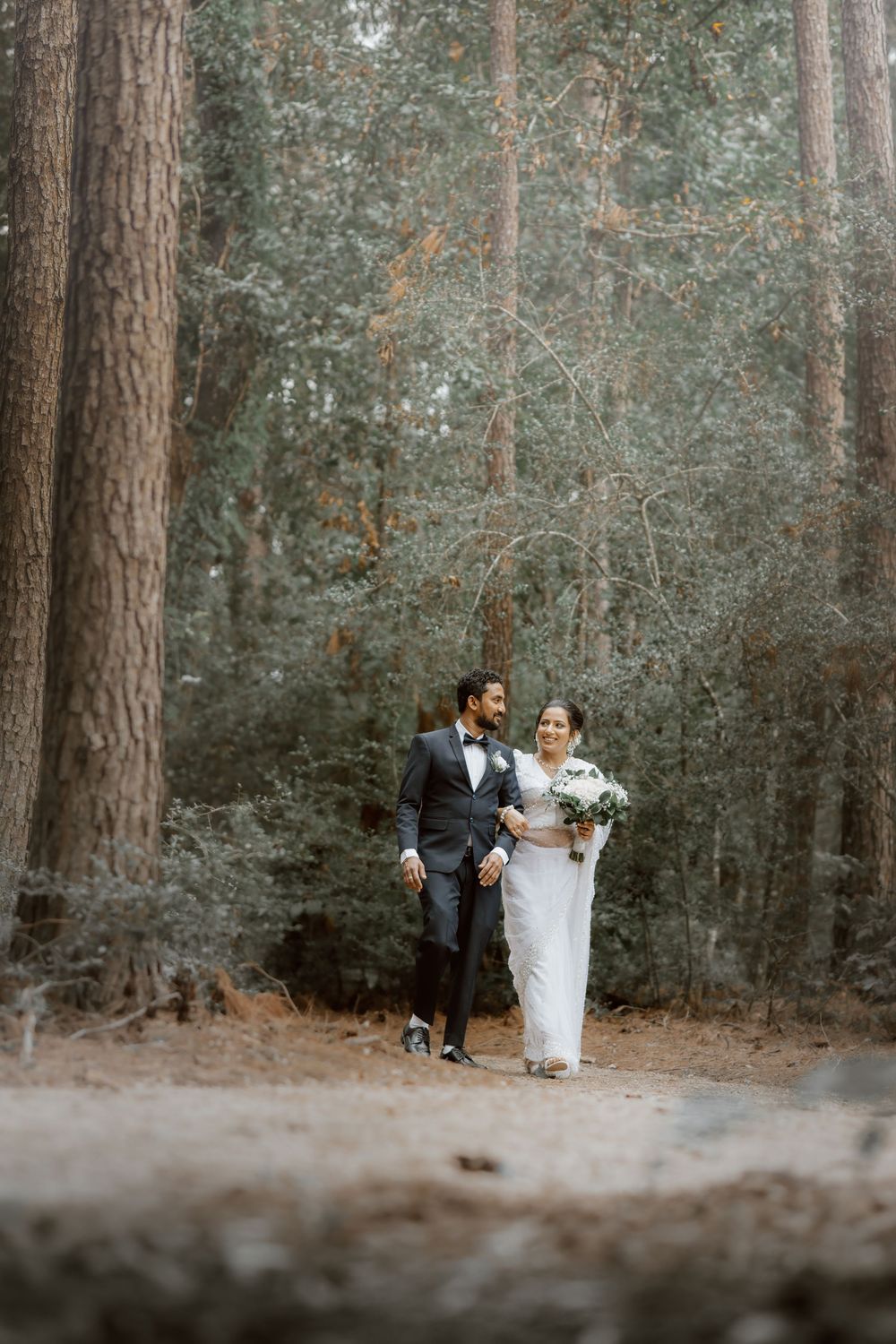 A romantic wedding couple stands together in a serene forest setting surrounded by tall pine trees.