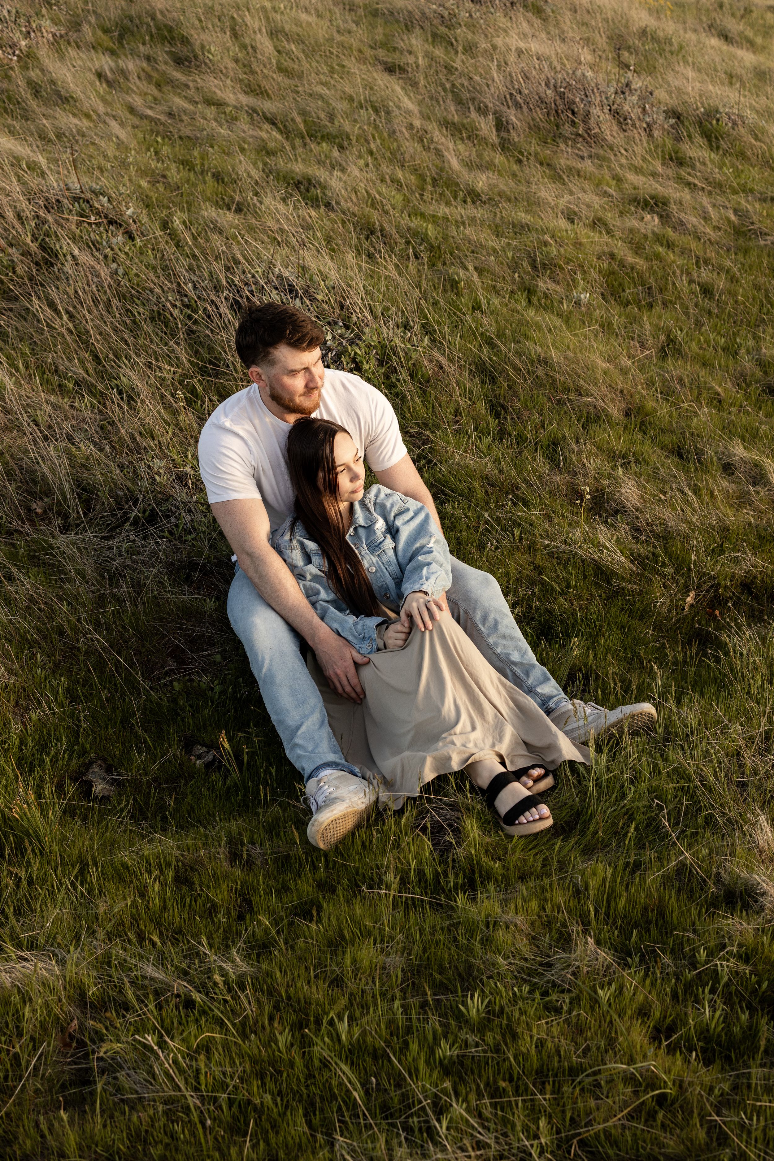 Couple sitting together on a grassy hillside, woman leaning against man in golden hour sunlight.