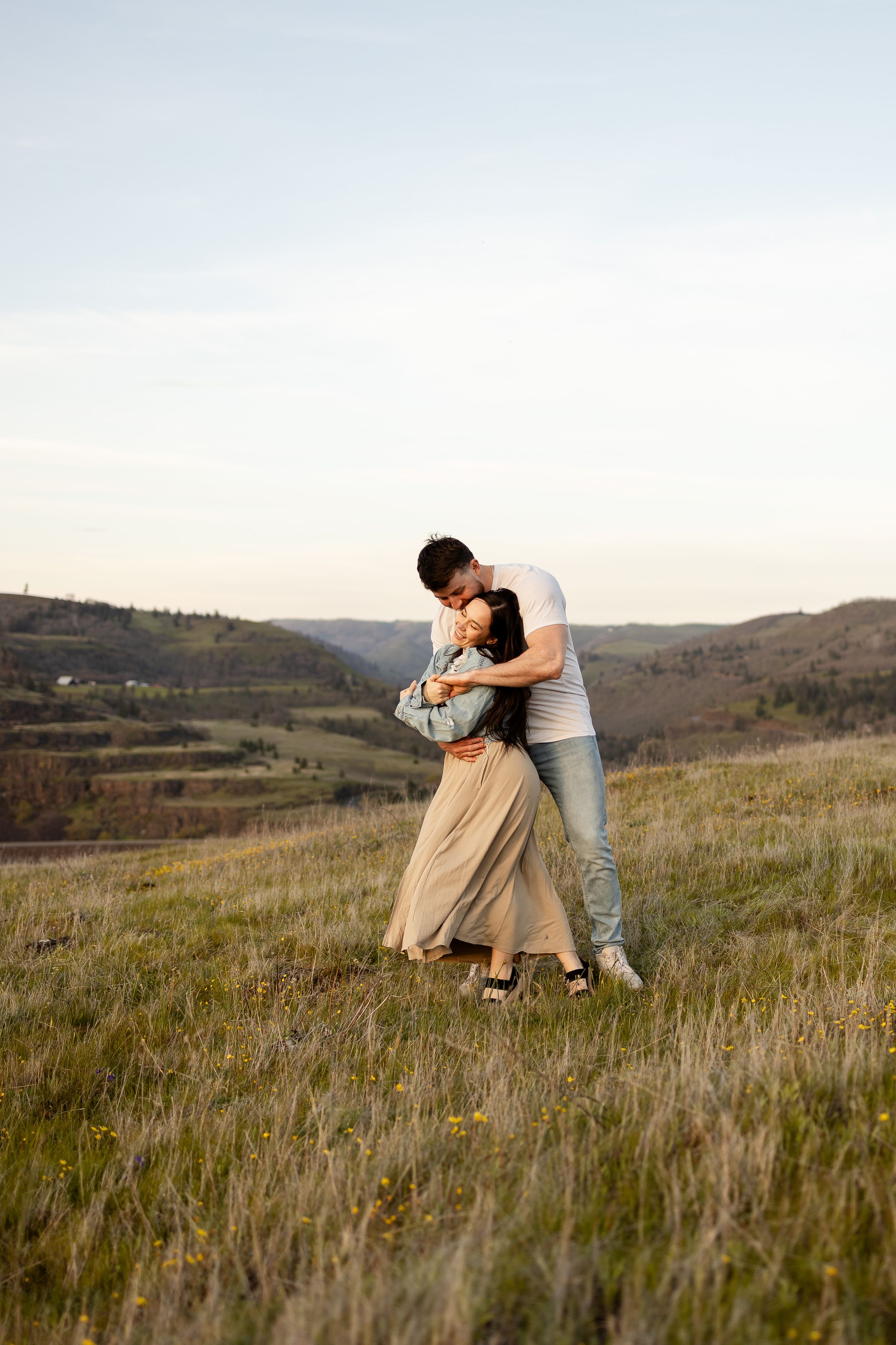 Couple embracing romantically on a green hillside with scenic valley and mountains in the background at dusk.
