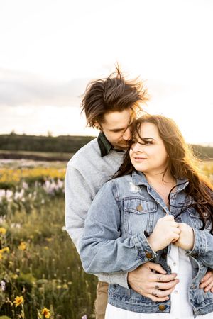 Couple embracing in a wildflower field at golden hour, woman in denim jacket, man nuzzling her hair.