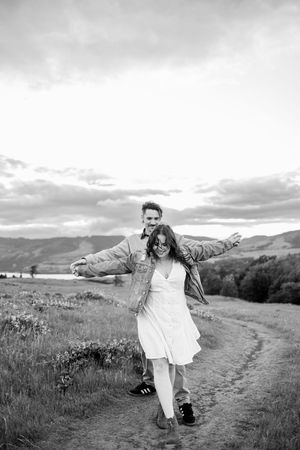 Black and white photo of a joyful couple on a rural path, woman in white dress with arms outstretched, mountains behind.