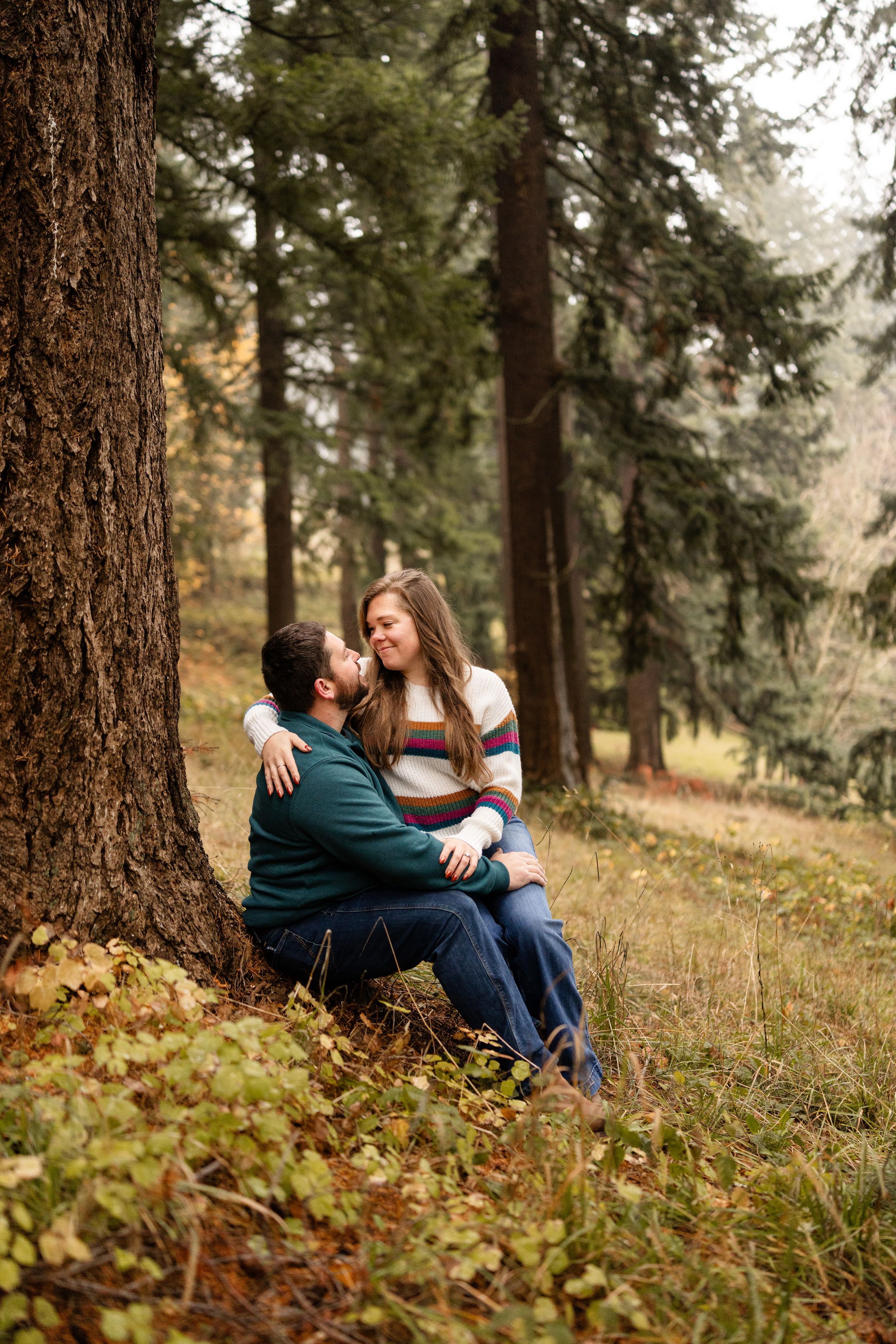 Couple sitting together against a tall tree in an autumn forest, woman in striped sweater embracing man in teal jacket.