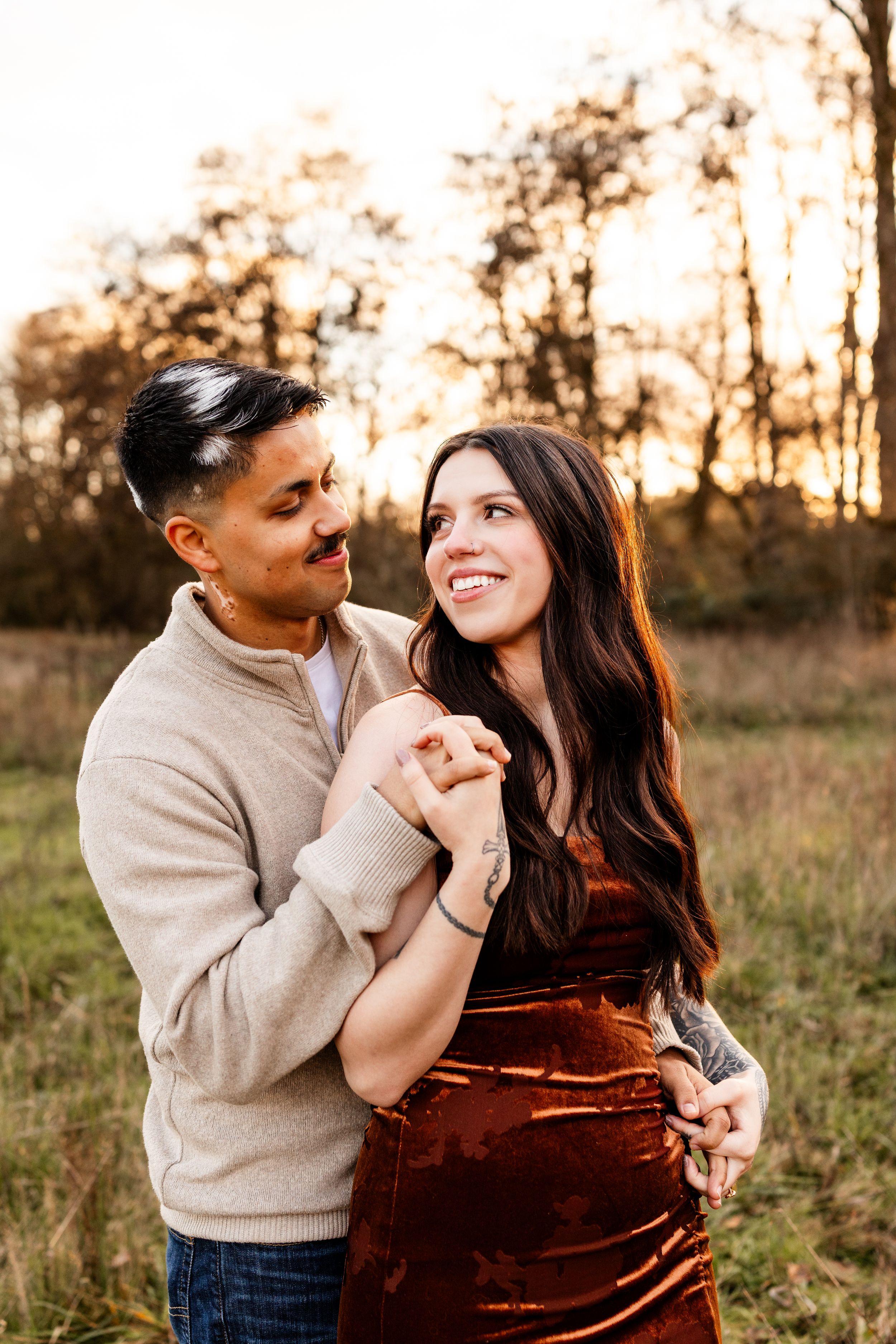 A couple embraces in a golden sunset field, the woman in a rust velvet dress smiling warmly as the man holds her hands.