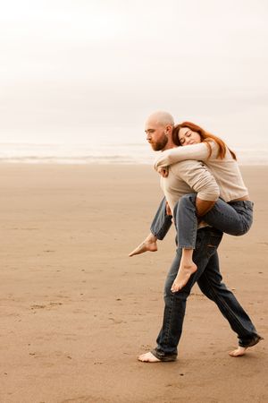 A bald man carries a redhead woman piggyback on a sandy beach, both wearing jeans and sweaters.