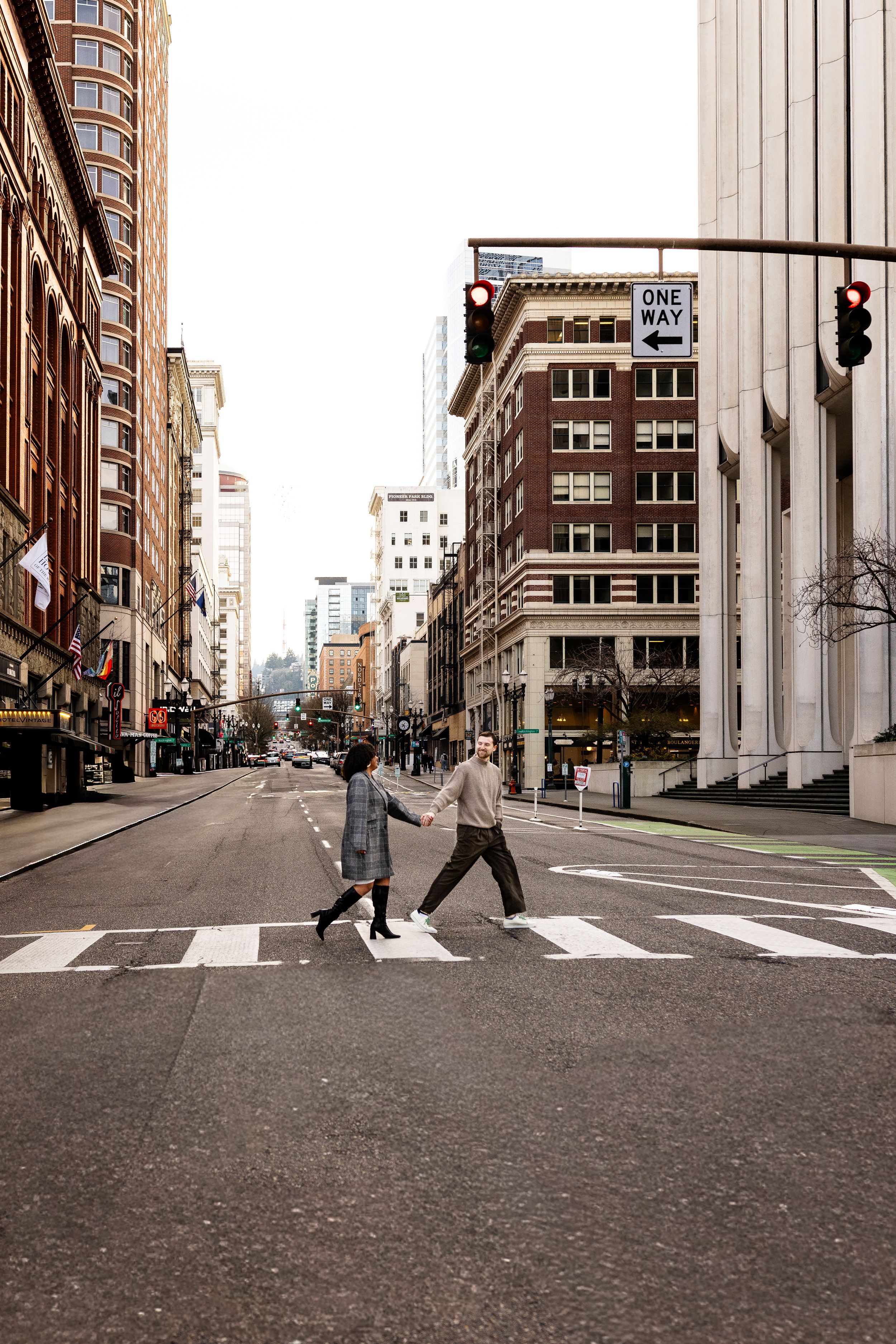 Couple holding hands crossing a zebra crosswalk in a busy urban city street with tall buildings and a one way sign.