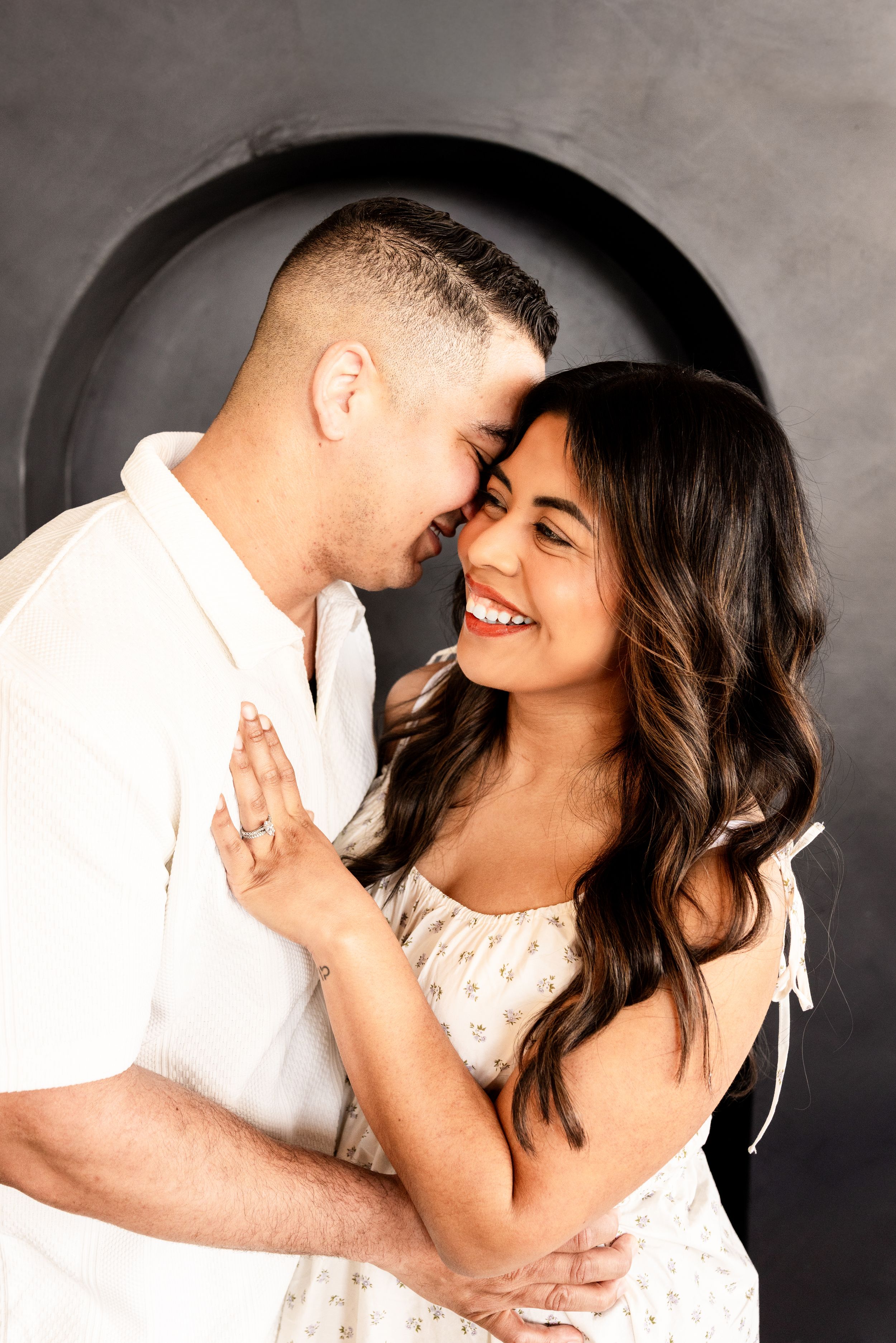 A smiling couple embraces tenderly in a studio, the woman in a floral dress and the man in white, sharing a romantic moment.
