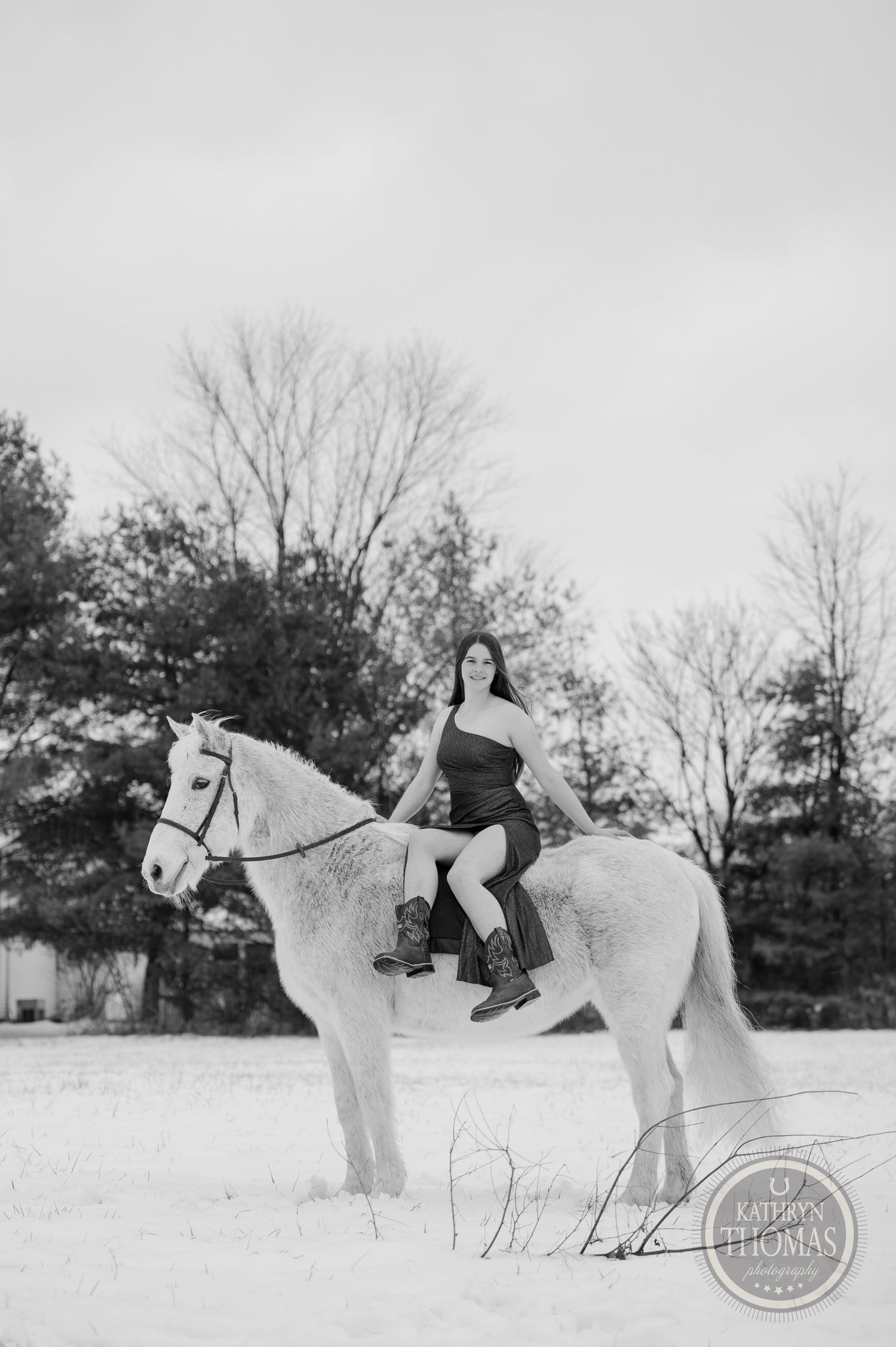 A person rides a white horse in a snowy field with bare winter trees in the background.