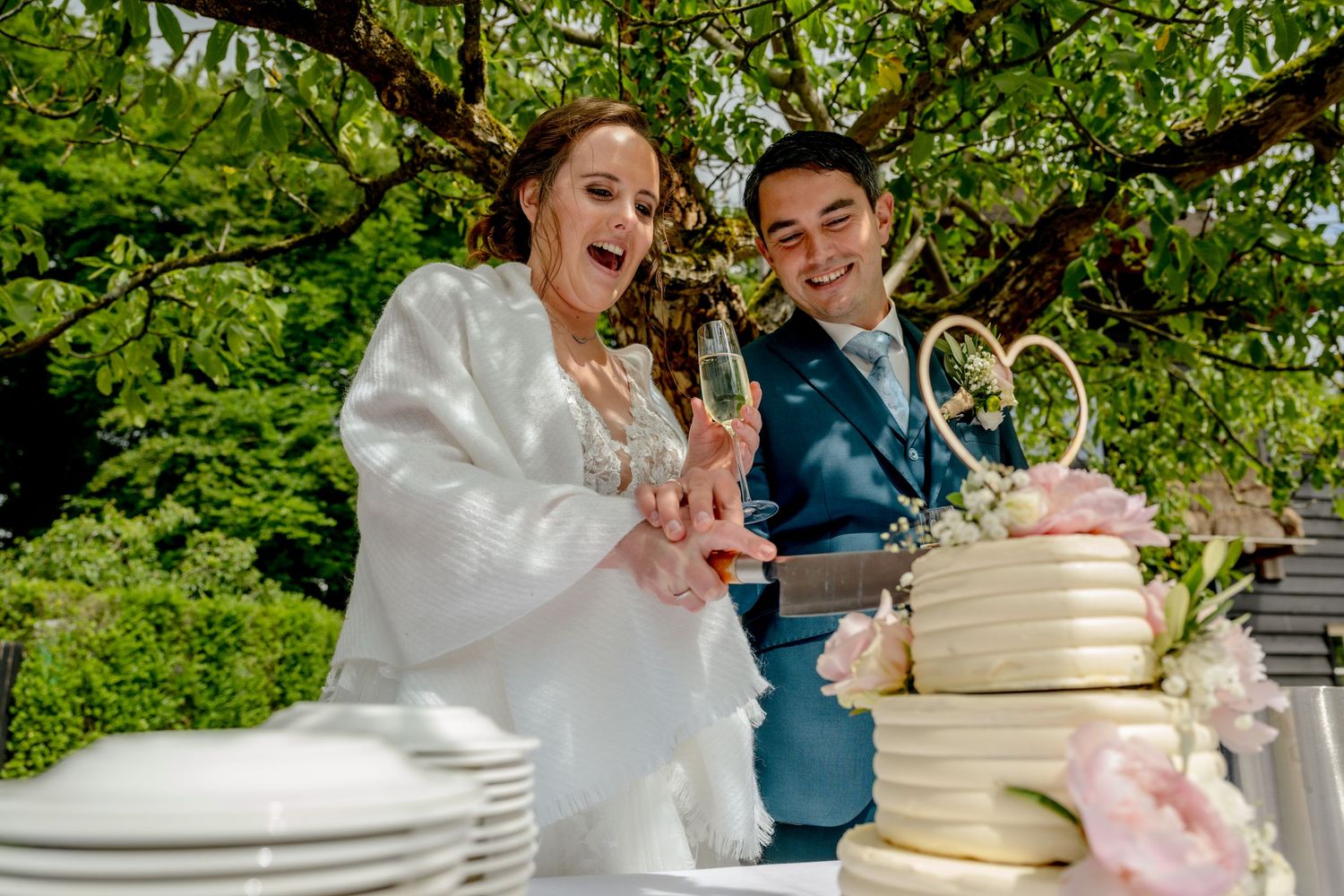 A couple shares a joyful moment while cutting their elegant white wedding cake decorated with pink flowers in a garden setting.