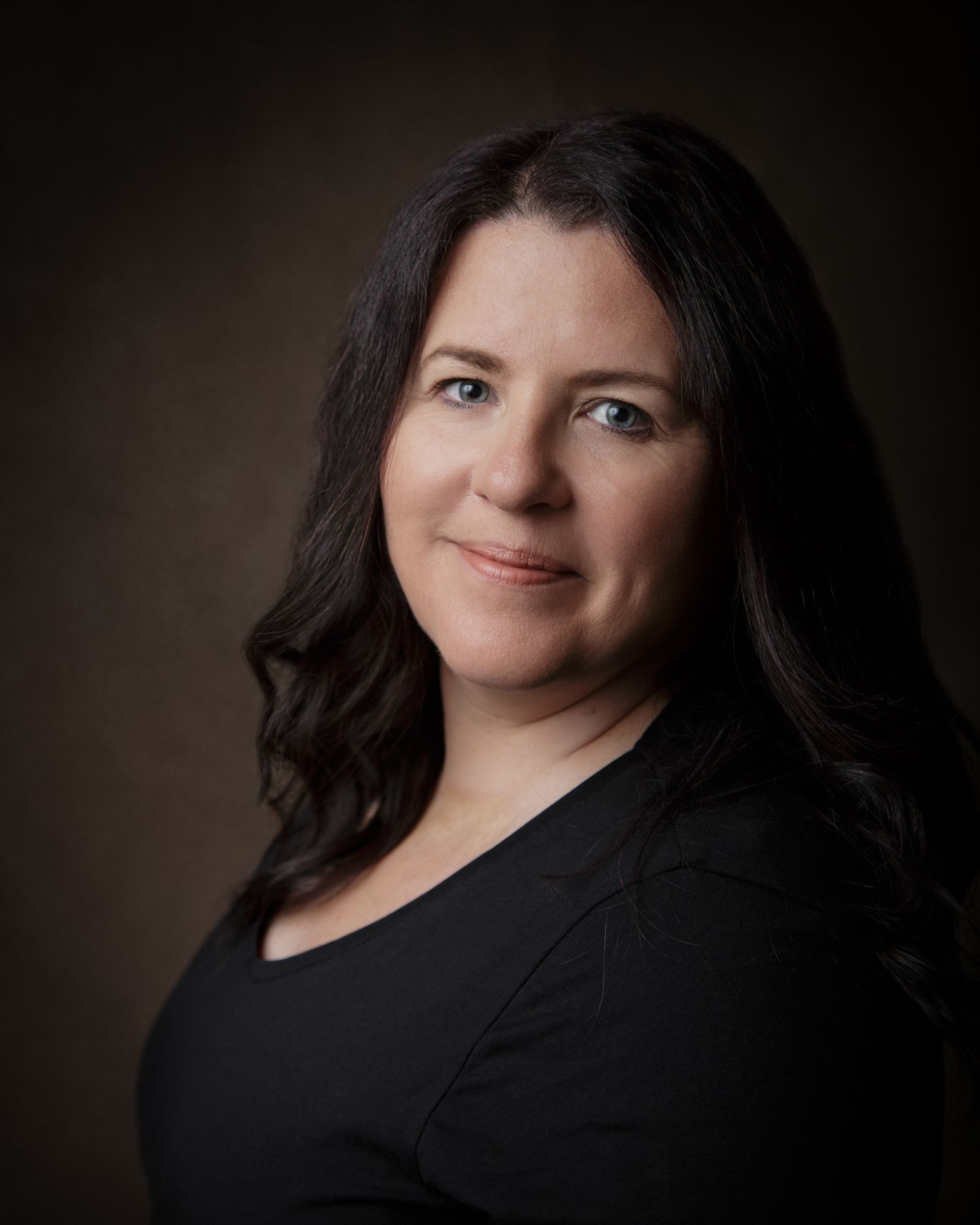A woman with long hair wearing a black shirt, standing confidently against a neutral background.