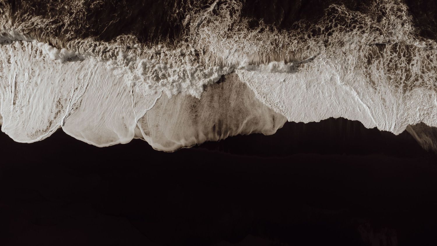 Golven die witte schuim achterlaten op een zwart strand vanuit de lucht gezien.