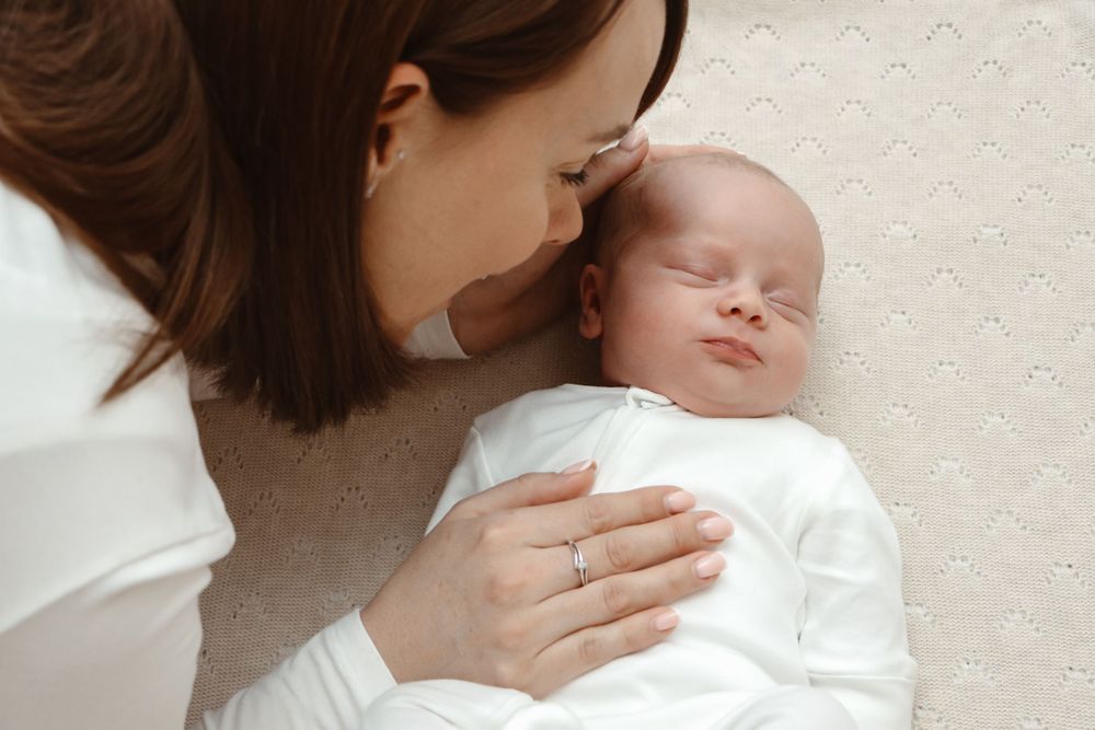 Mother lying next to sleeping newborn baby wrapped in white clothing.