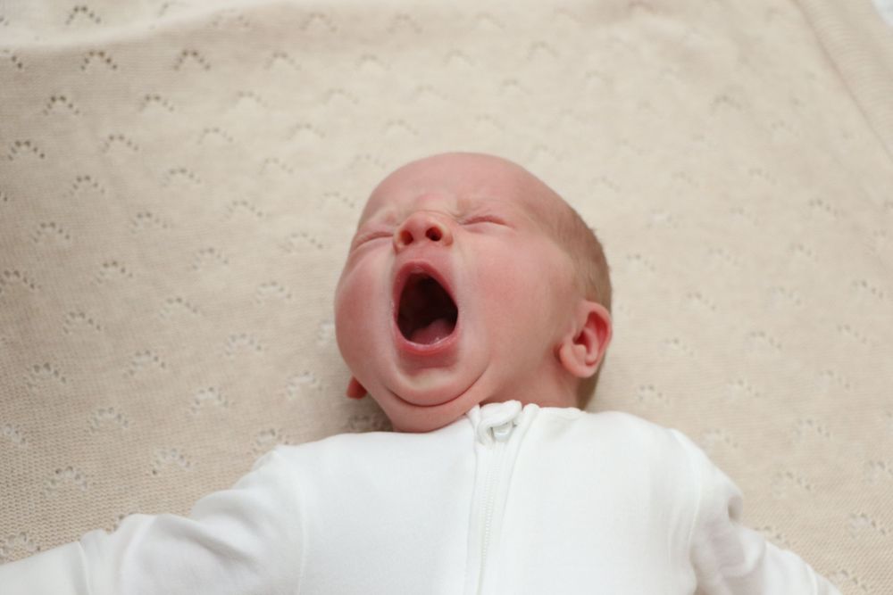 Newborn baby in white clothing yawning while lying on textured fabric.