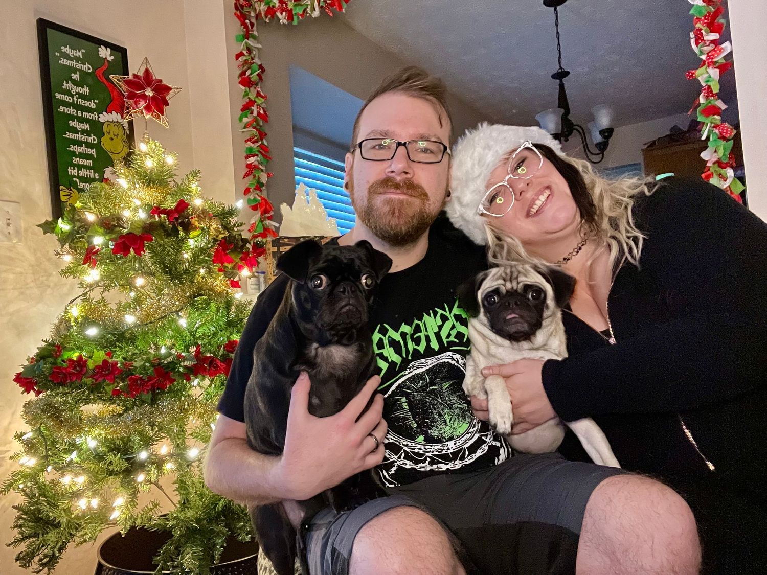 Couple holding black and pug dogs in front of Christmas tree with lights and decorations.