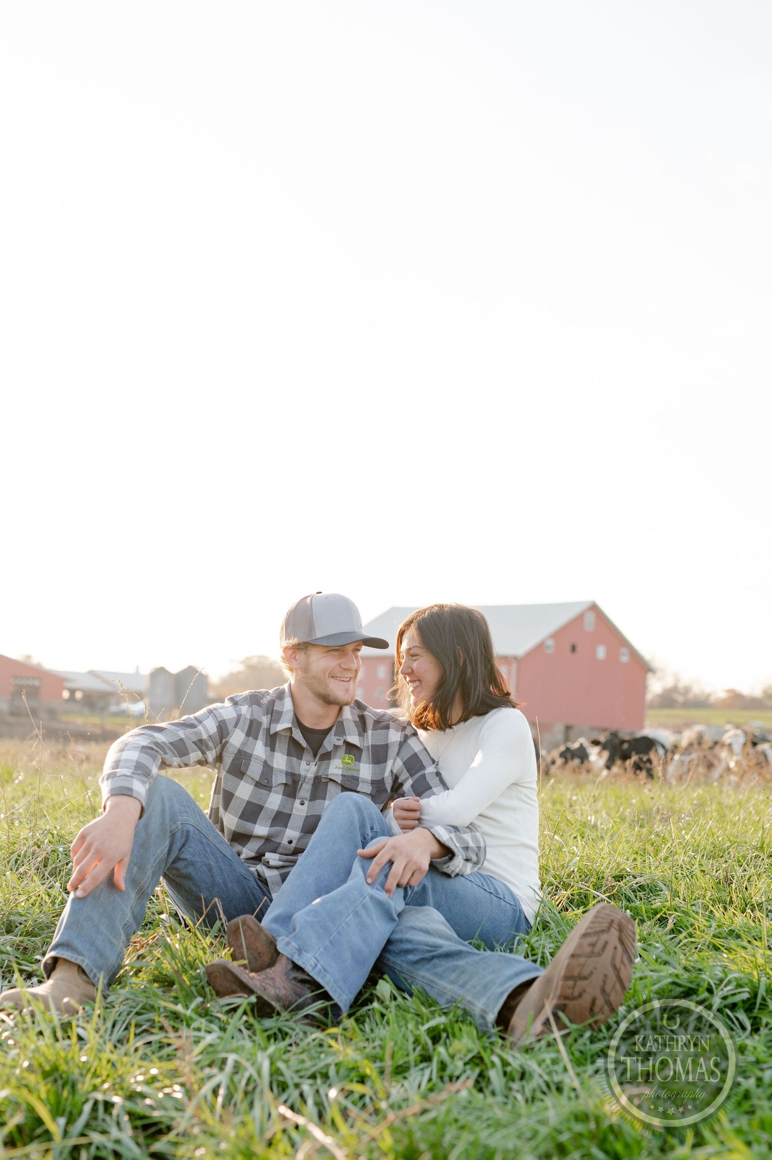 A couple sits together in a sunlit field with a pastoral farm setting in the background.