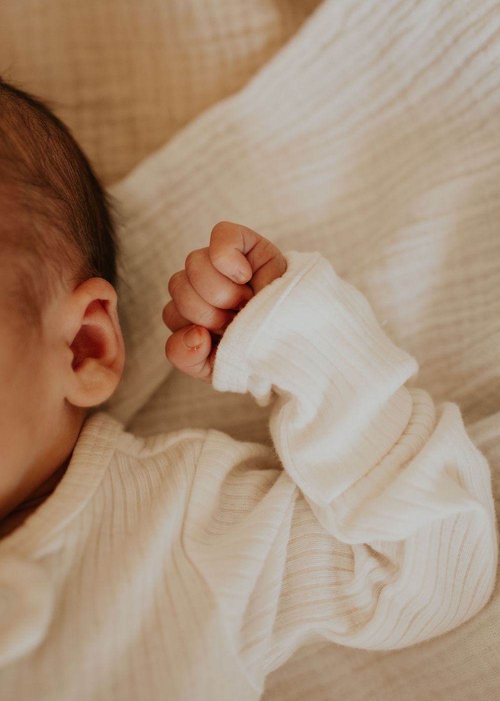 Close-up of a tiny baby hand in white sleeve against soft white bedding while sleeping.