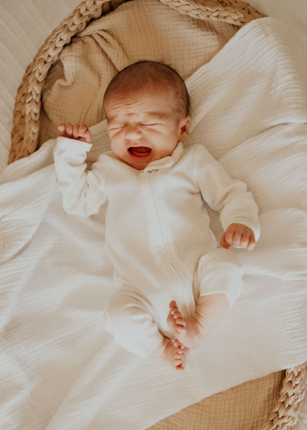 A newborn baby in a white onesie yawns while lying on soft white bedding in a woven bassinet.