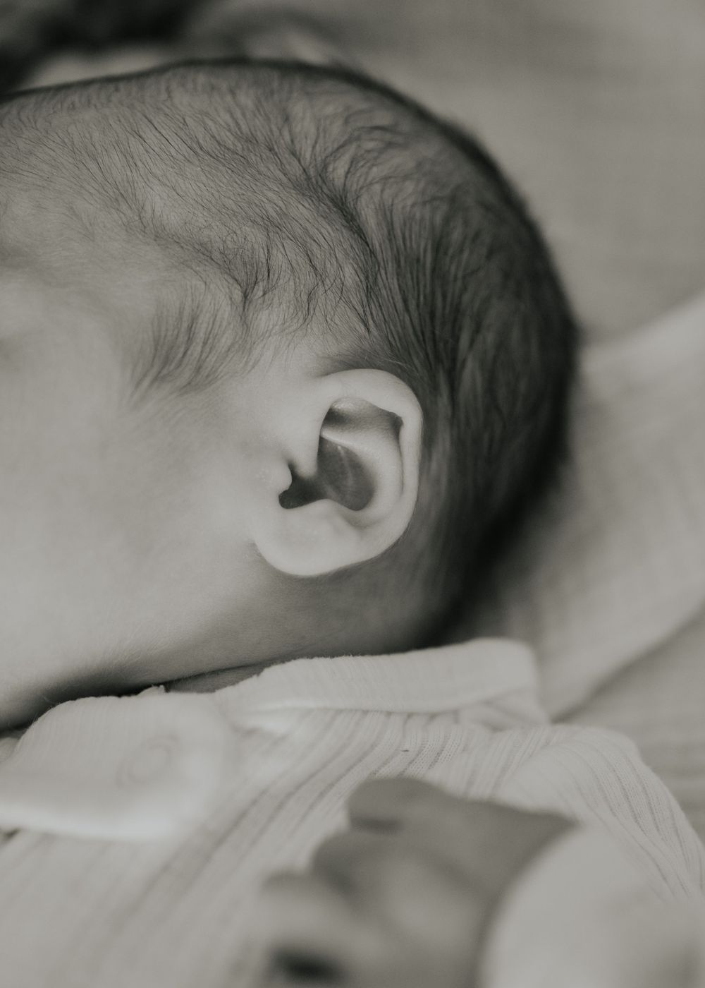 Close-up black and white photograph of a baby's head showing tiny ear and dark hair against soft bedding.