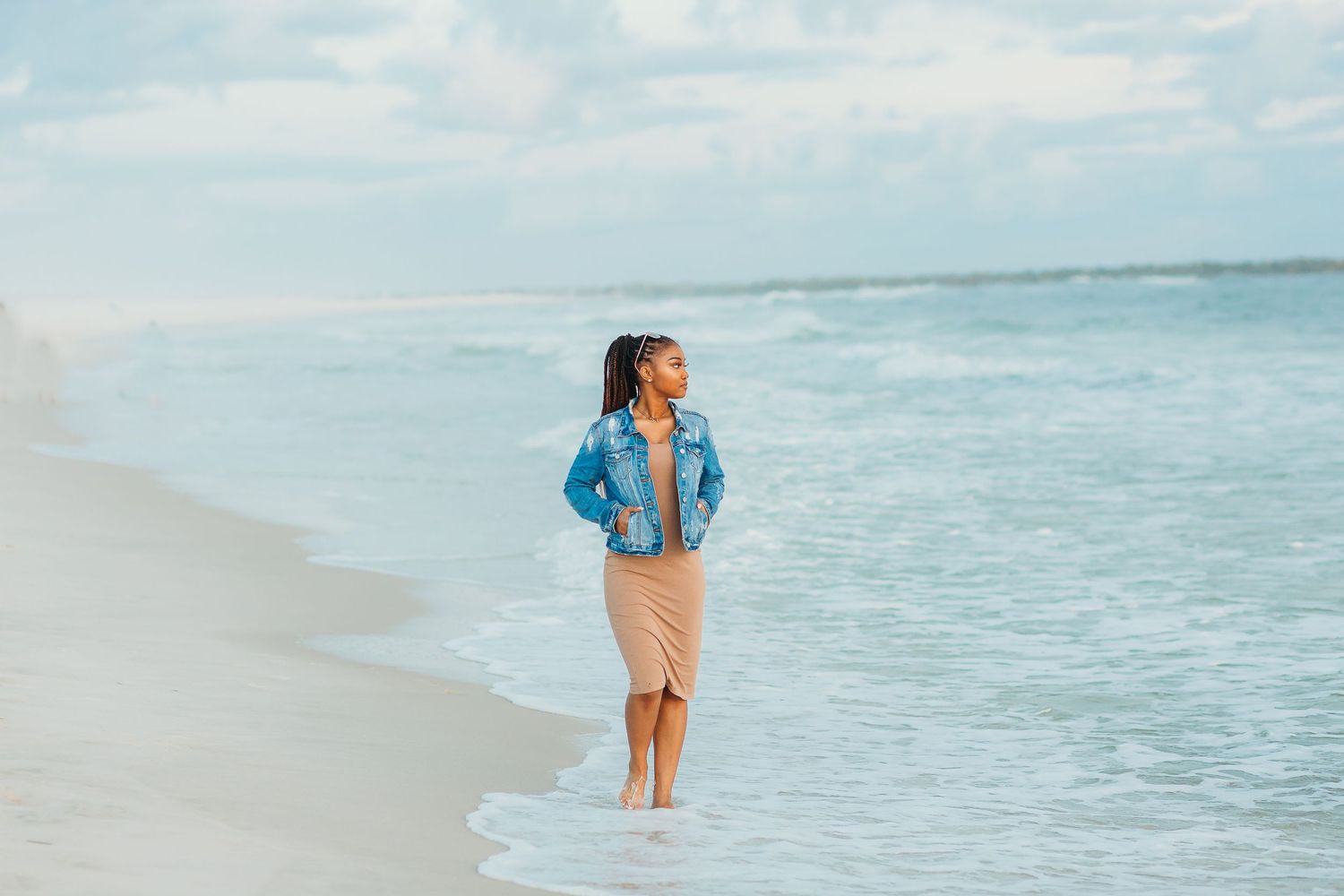 A person in a denim jacket and pink pants strolls peacefully along a serene beach with turquoise waters.