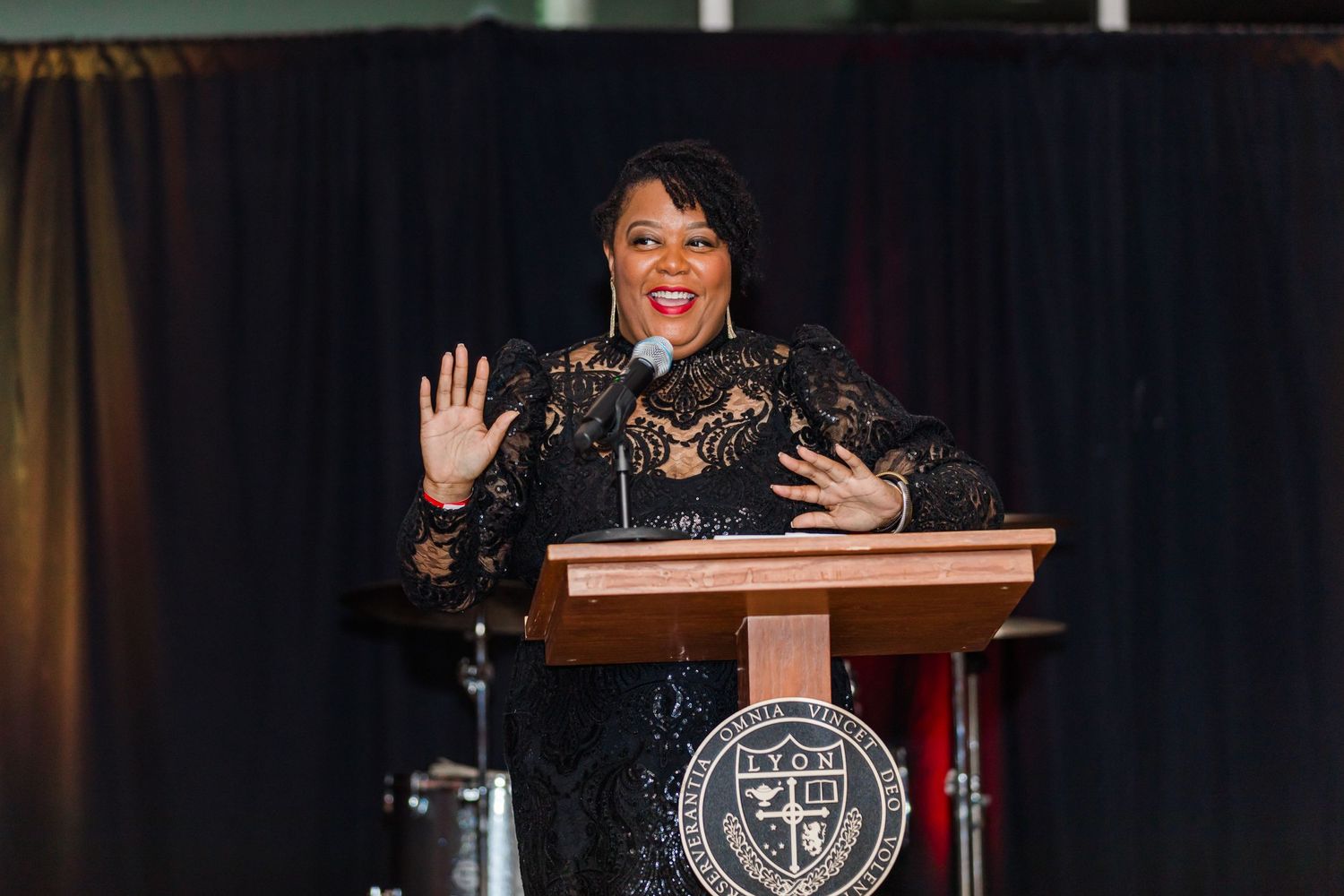 A speaker wearing black lace stands at a podium with a university seal, gesturing warmly to the audience.