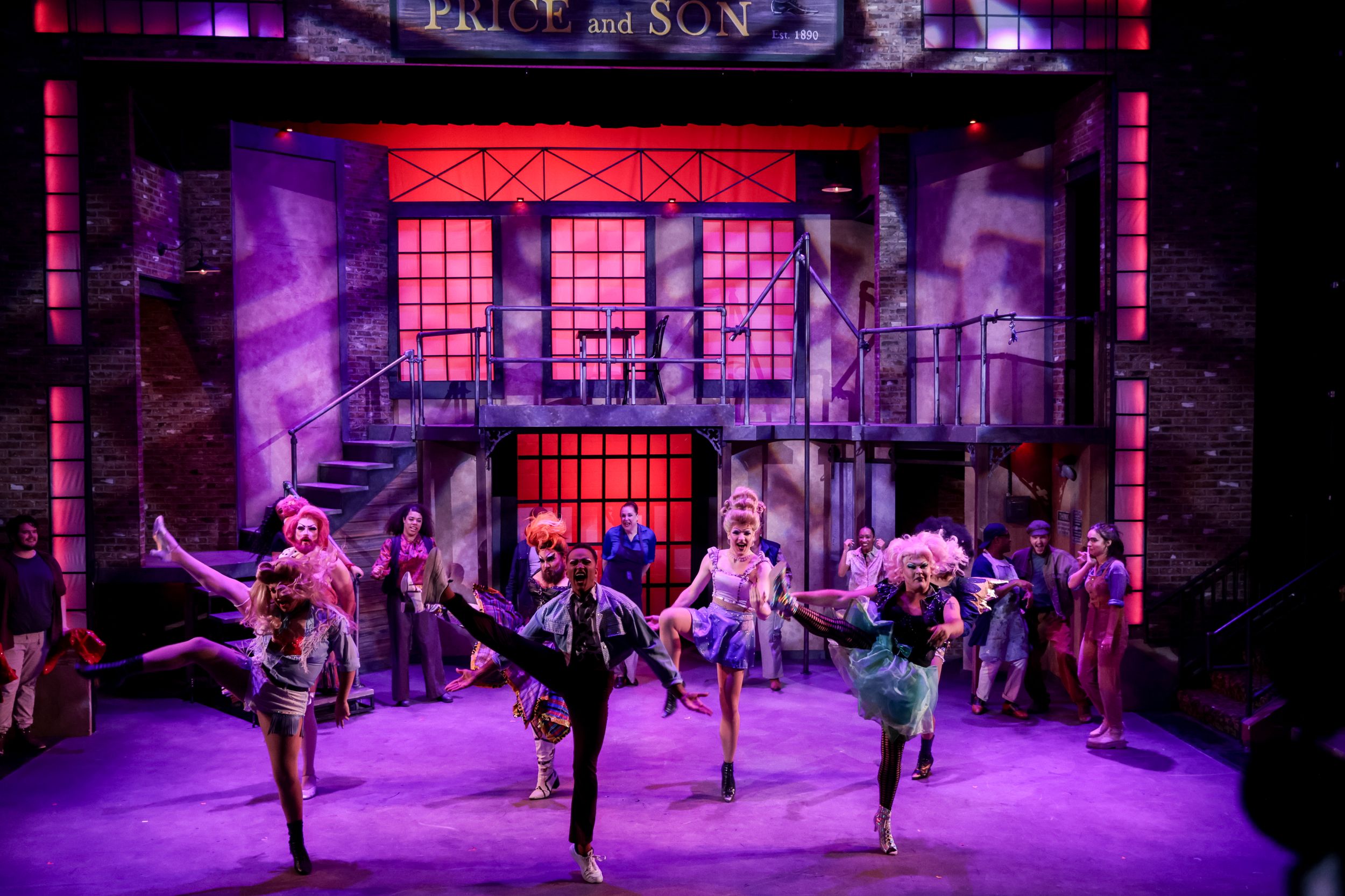 Dancers perform an energetic dance number on a theatrical stage with purple lighting and architectural set pieces. Theatre photography.