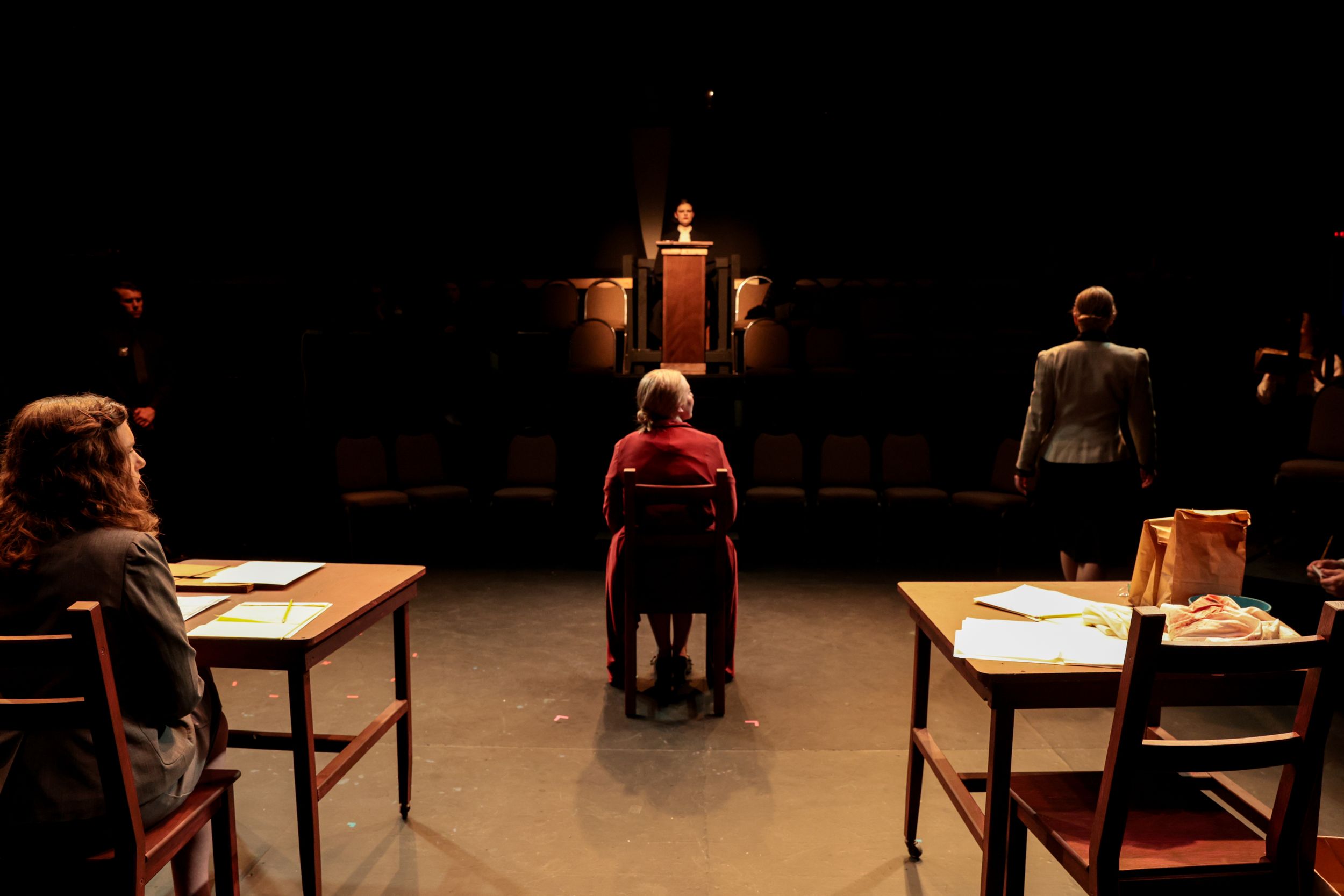 Dramatic theater scene with performers seated at tables in a dimly lit institutional setting. Theatre photography.