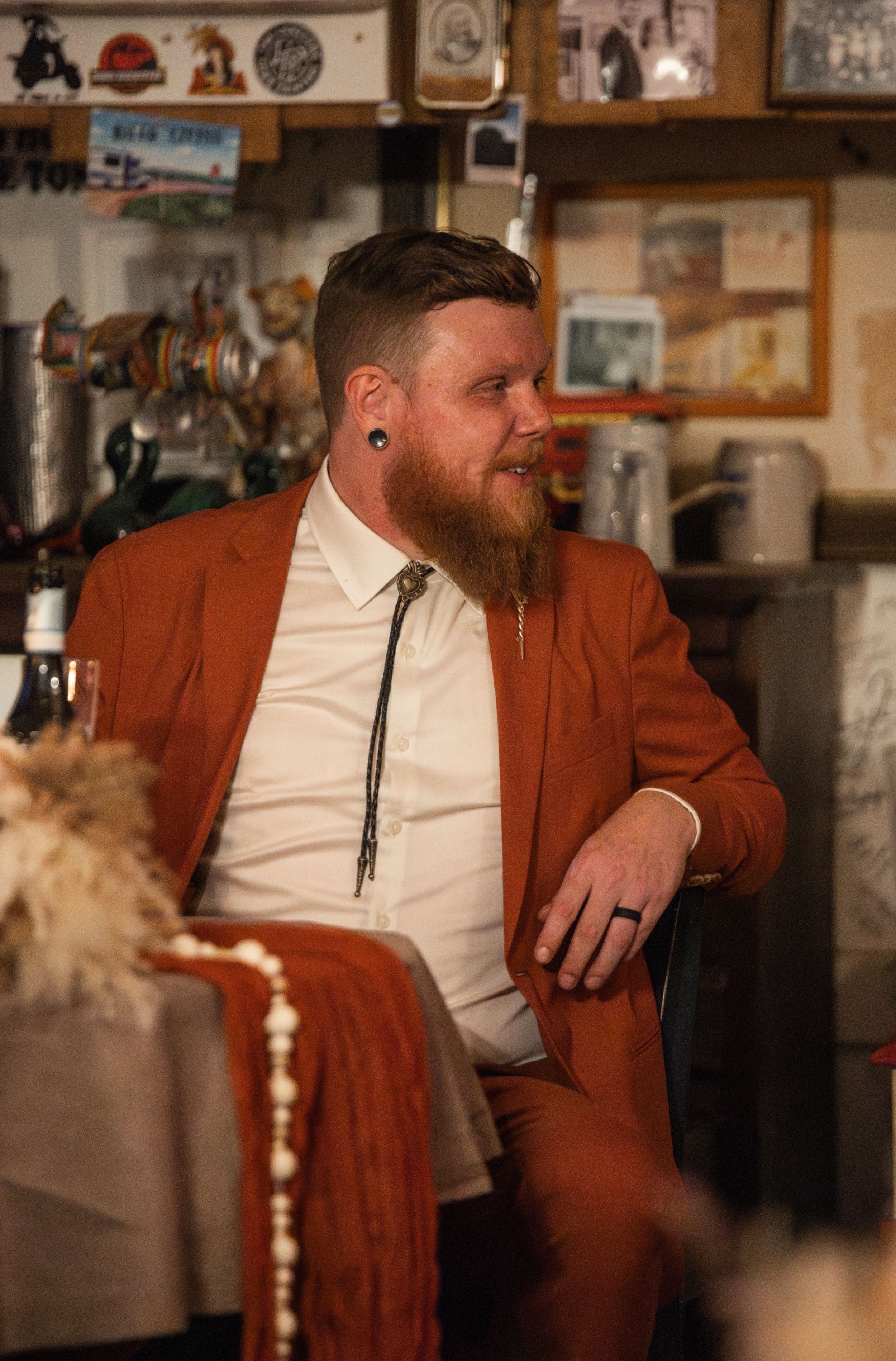 A man in a rust-colored suit sits casually in a room filled with vintage decor and memorabilia.
