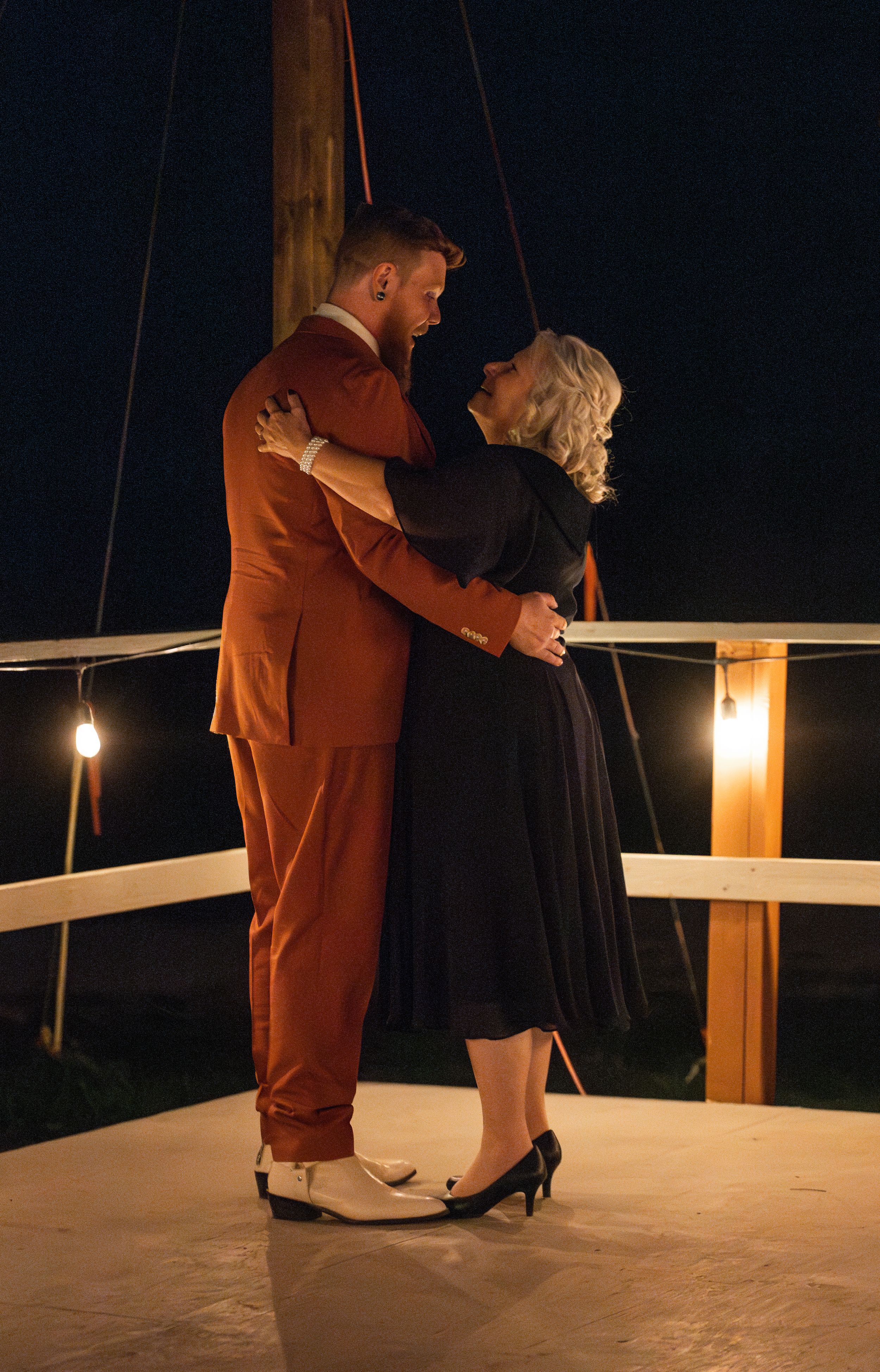 A couple dances on a lit deck at night, with the groom in rust suit and partner in black dress.