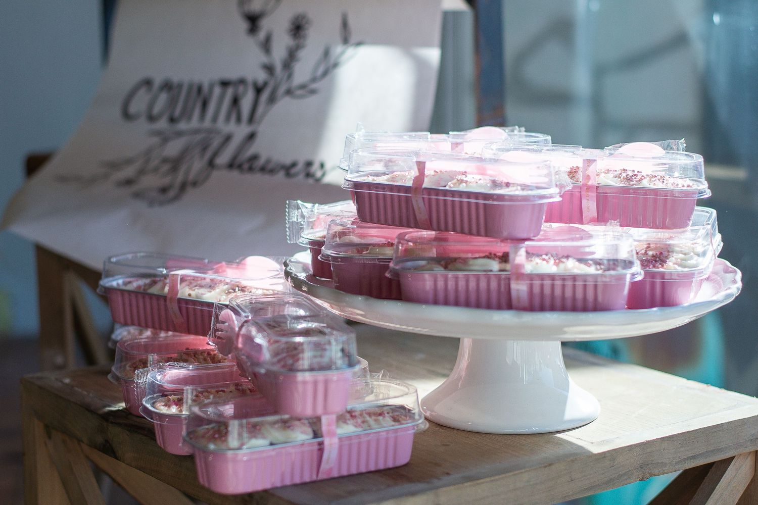 Pink bakery boxes with clear lids filled with cupcakes displayed on a wooden table and white cake stand.
