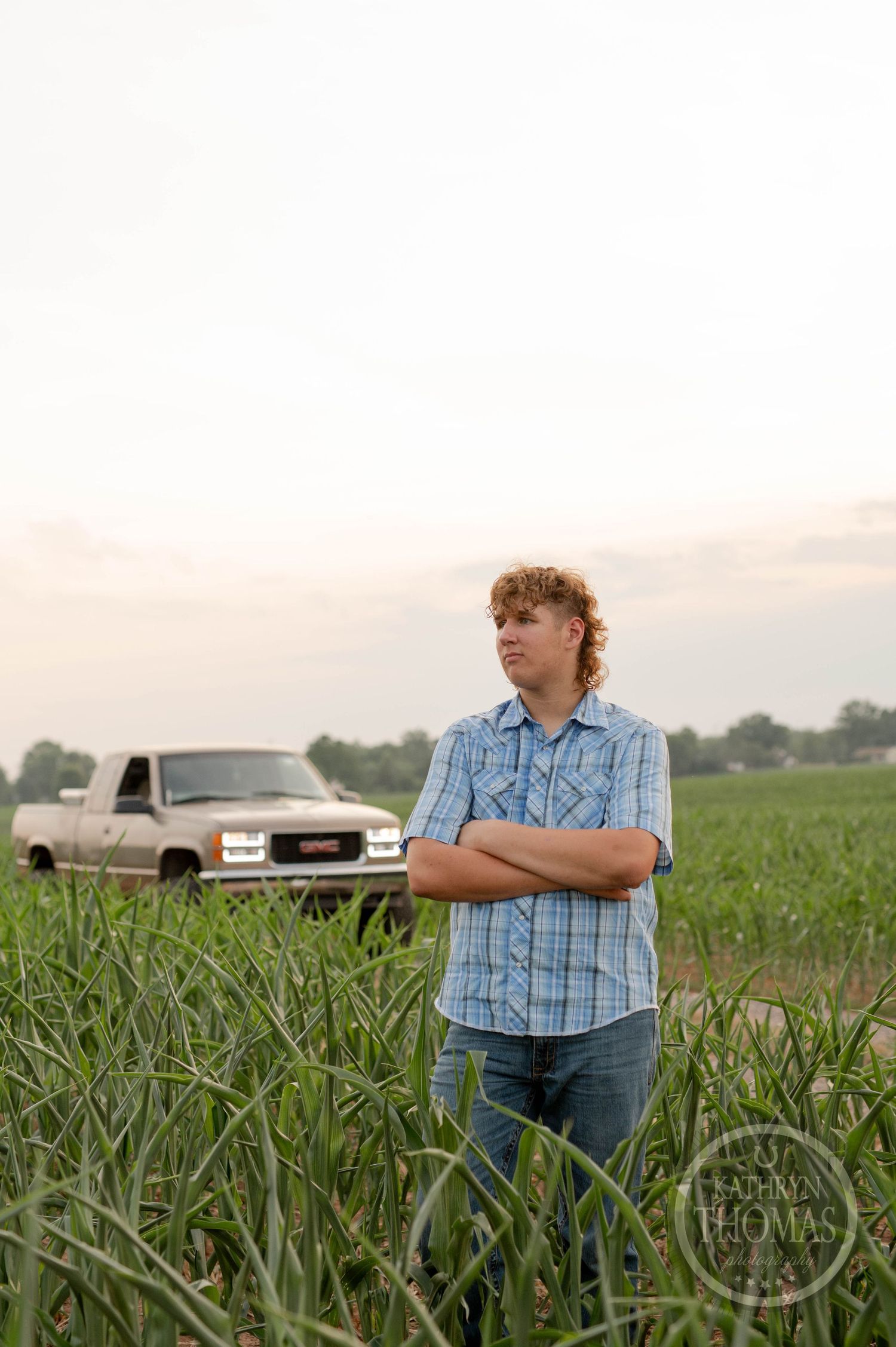 Person in plaid shirt standing in cornfield with lifted GMC truck parked on dirt road.