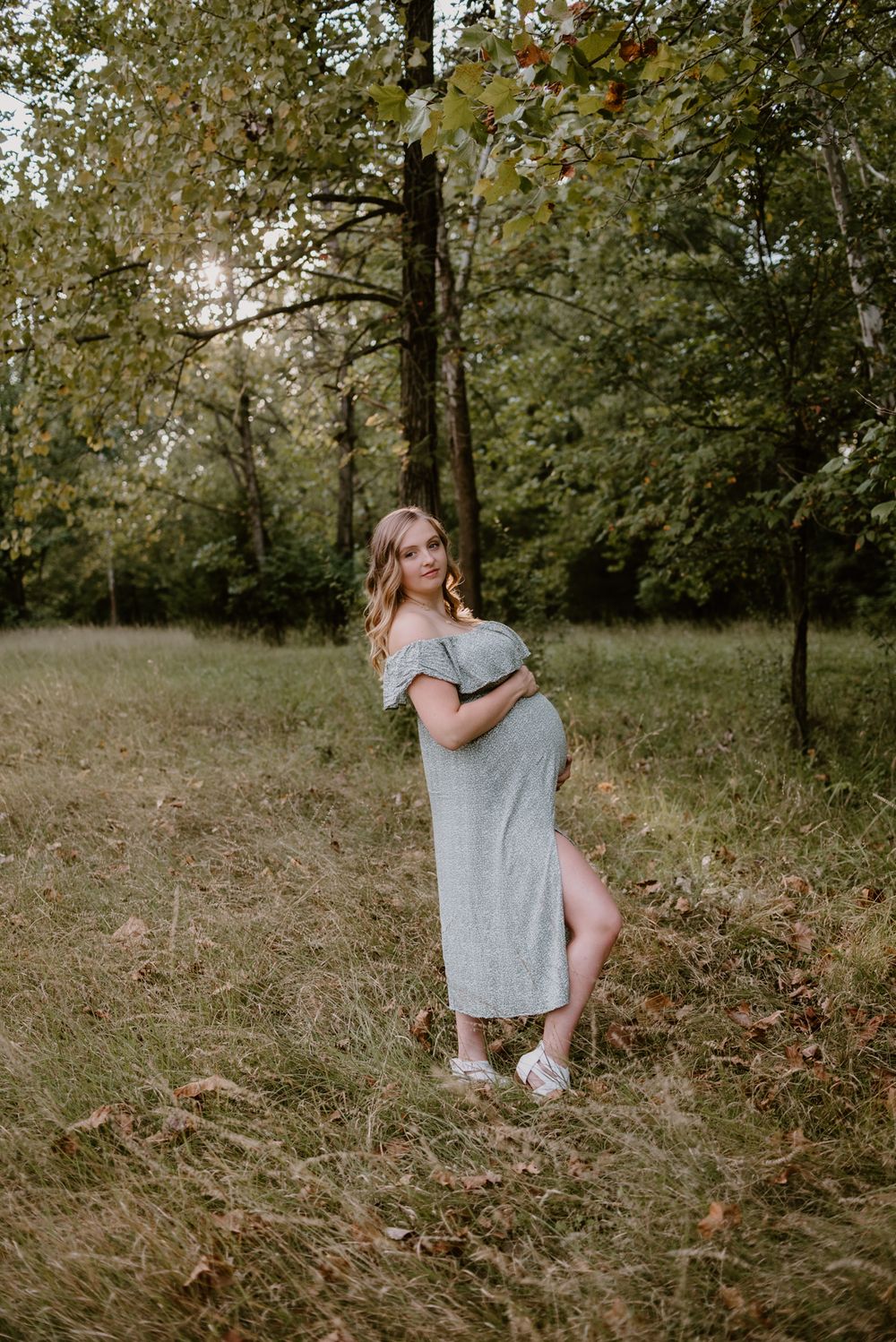 An expectant mother in a grey dress poses in a rustic woodland setting.