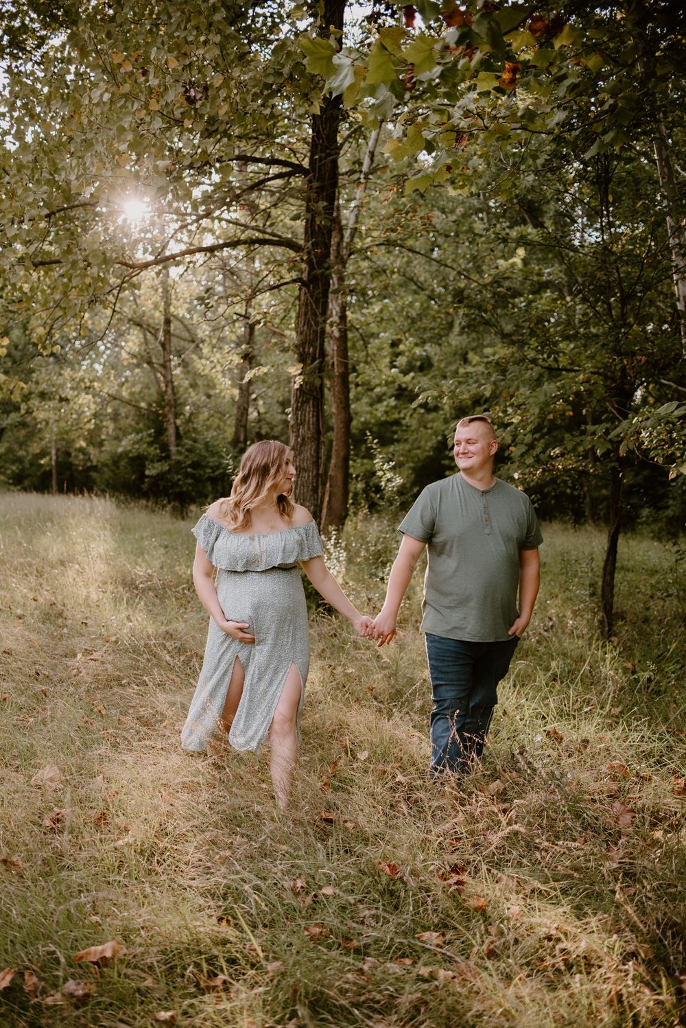 A couple holding hands while walking through a sunlit forest clearing during their maternity session.