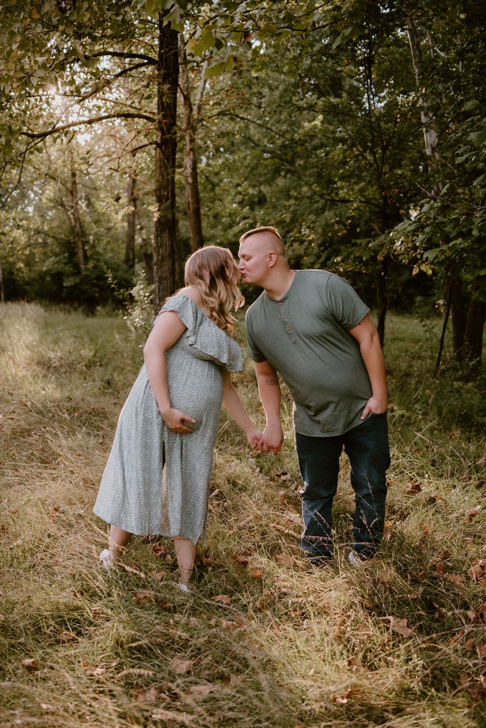 A couple shares an intimate moment during their maternity photoshoot in a wooded area.