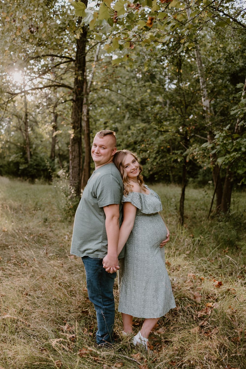 A couple stands back-to-back in a sunlit wooded area during a maternity photoshoot.
