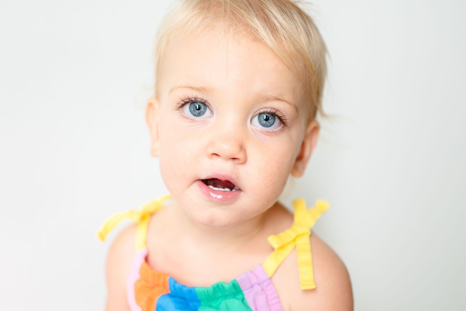 A baby with bright blue eyes wearing a colorful rainbow dress with yellow bow straps against a white background.