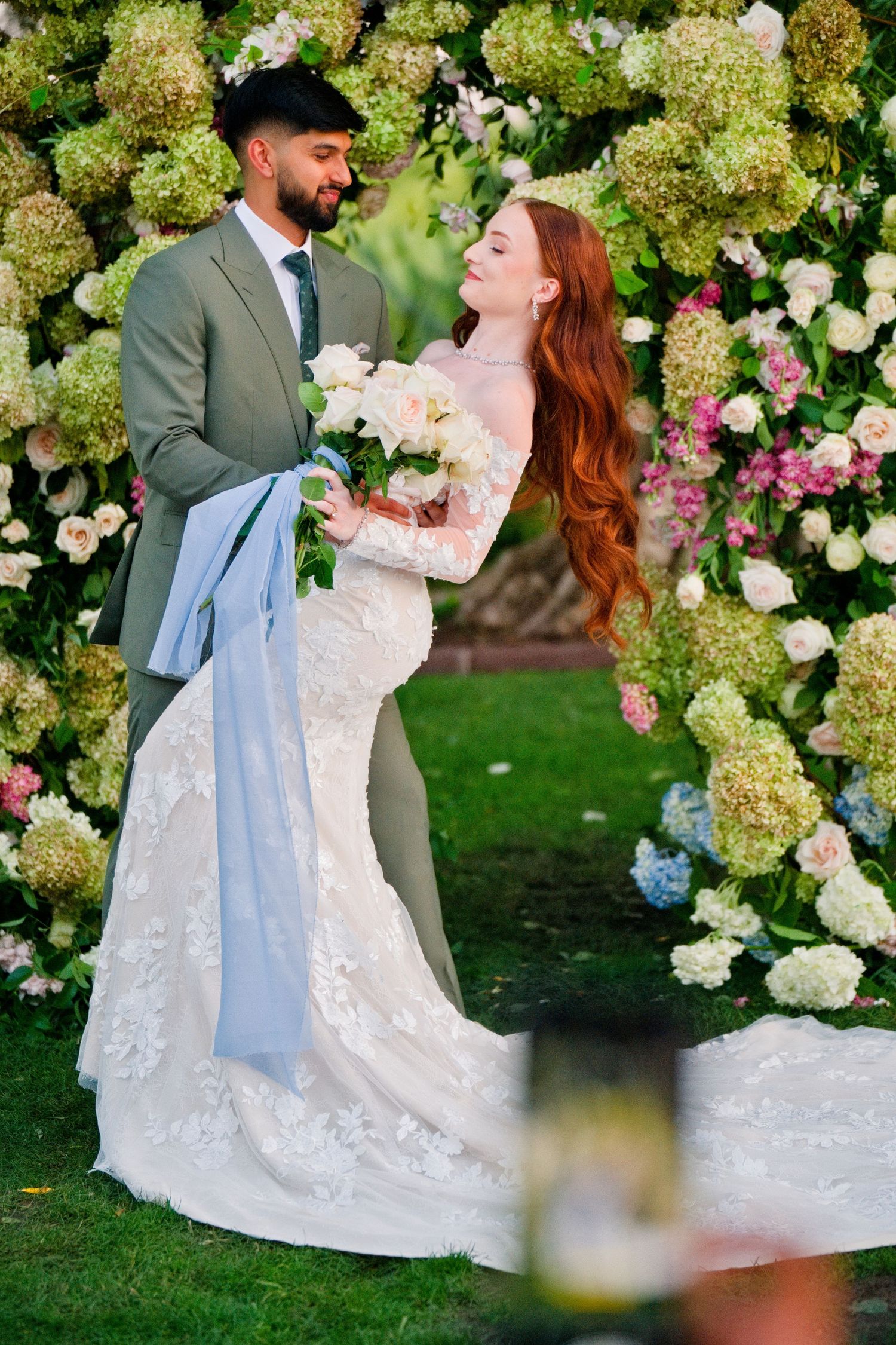 A bride in a lace wedding gown and groom in a gray suit embrace in front of a floral arch with hydrangeas and roses.