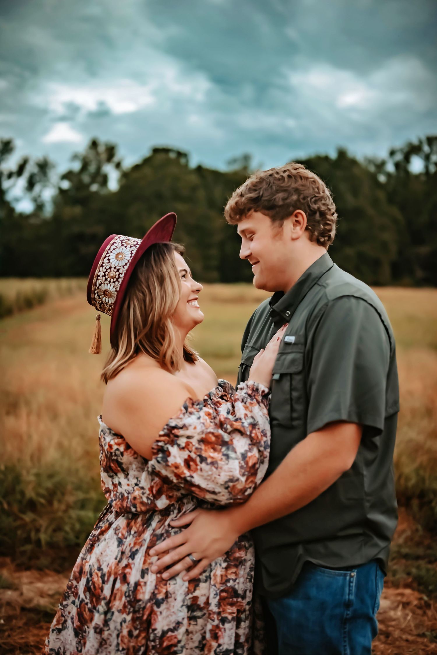 Couple embraces in autumn field under moody sky as woman in floral dress and hat smiles joyfully.