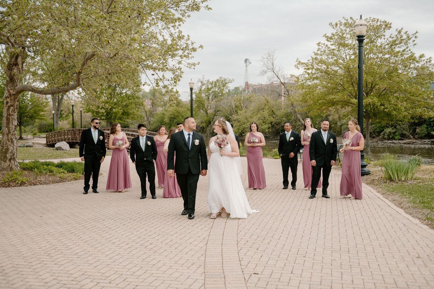 Wedding party in pink and teal attire poses together on wooden bridge over water during outdoor celebration.