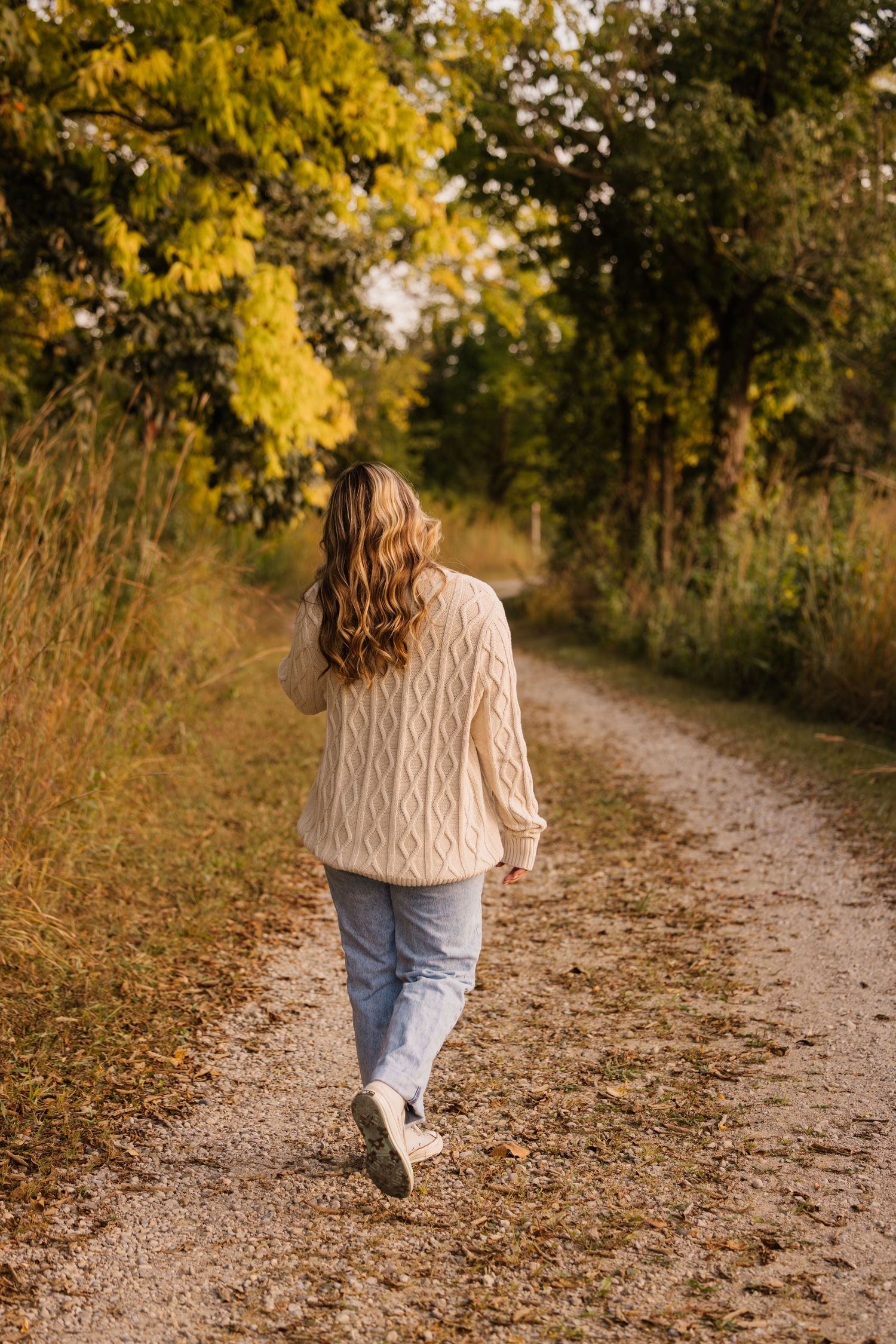 Person wearing a cozy cream cable knit sweater walks along an autumn path surrounded by golden foliage.