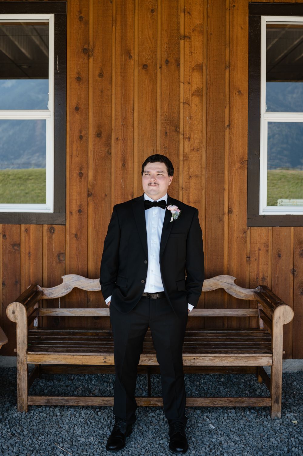 A groom in a black tuxedo and bowtie stands in front of a wooden wall between two windows on a rustic wooden bench.