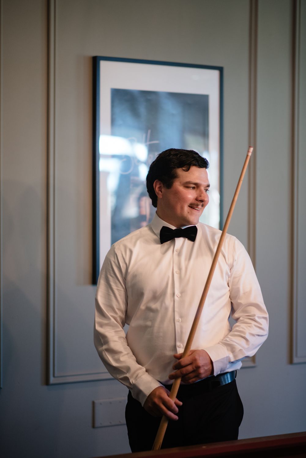 A billiards player in formal attire with bow tie and white shirt smiles while holding a pool cue in an elegant room.