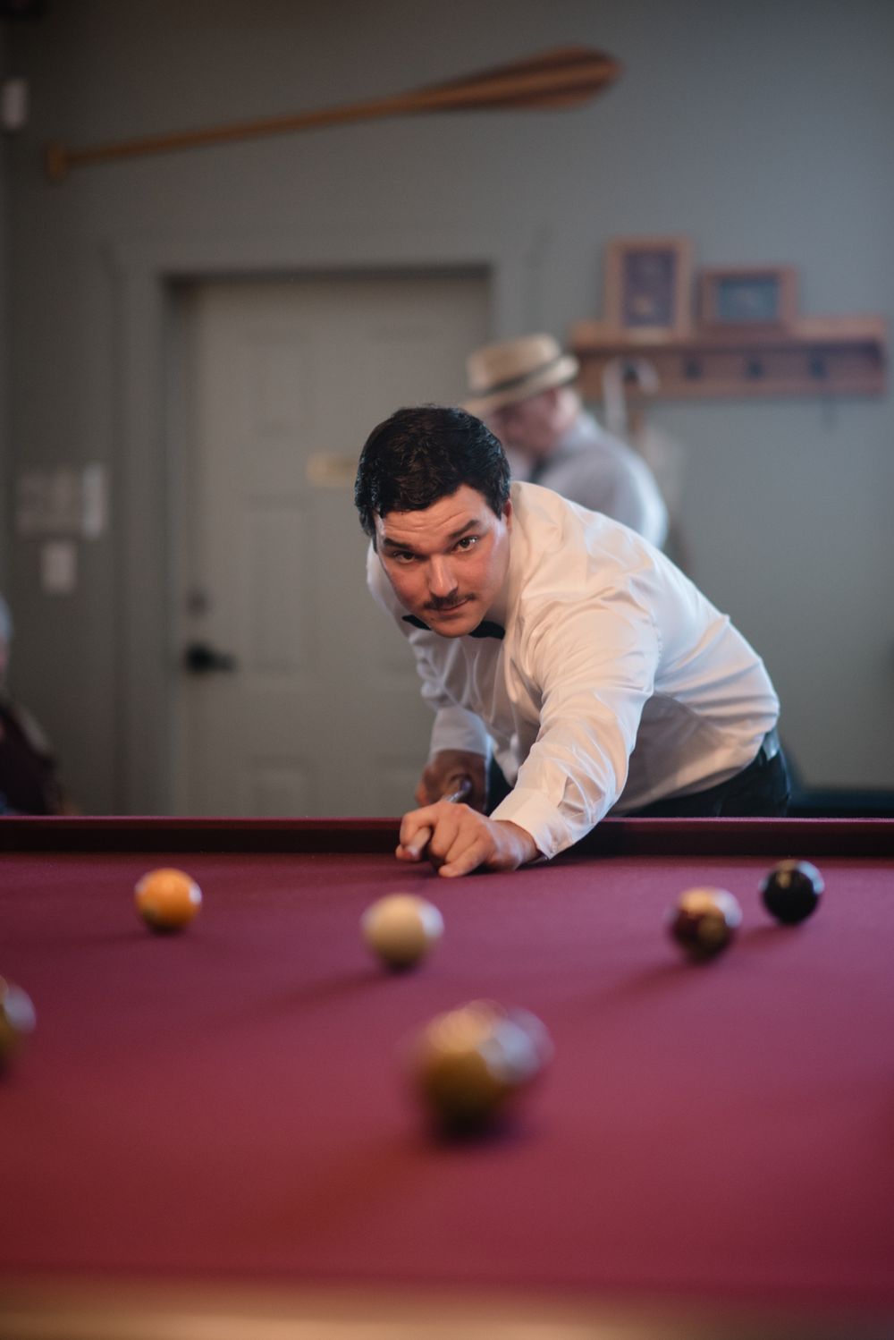 A person in a white shirt leans over a red pool table to take a shot while another person stands in the background.