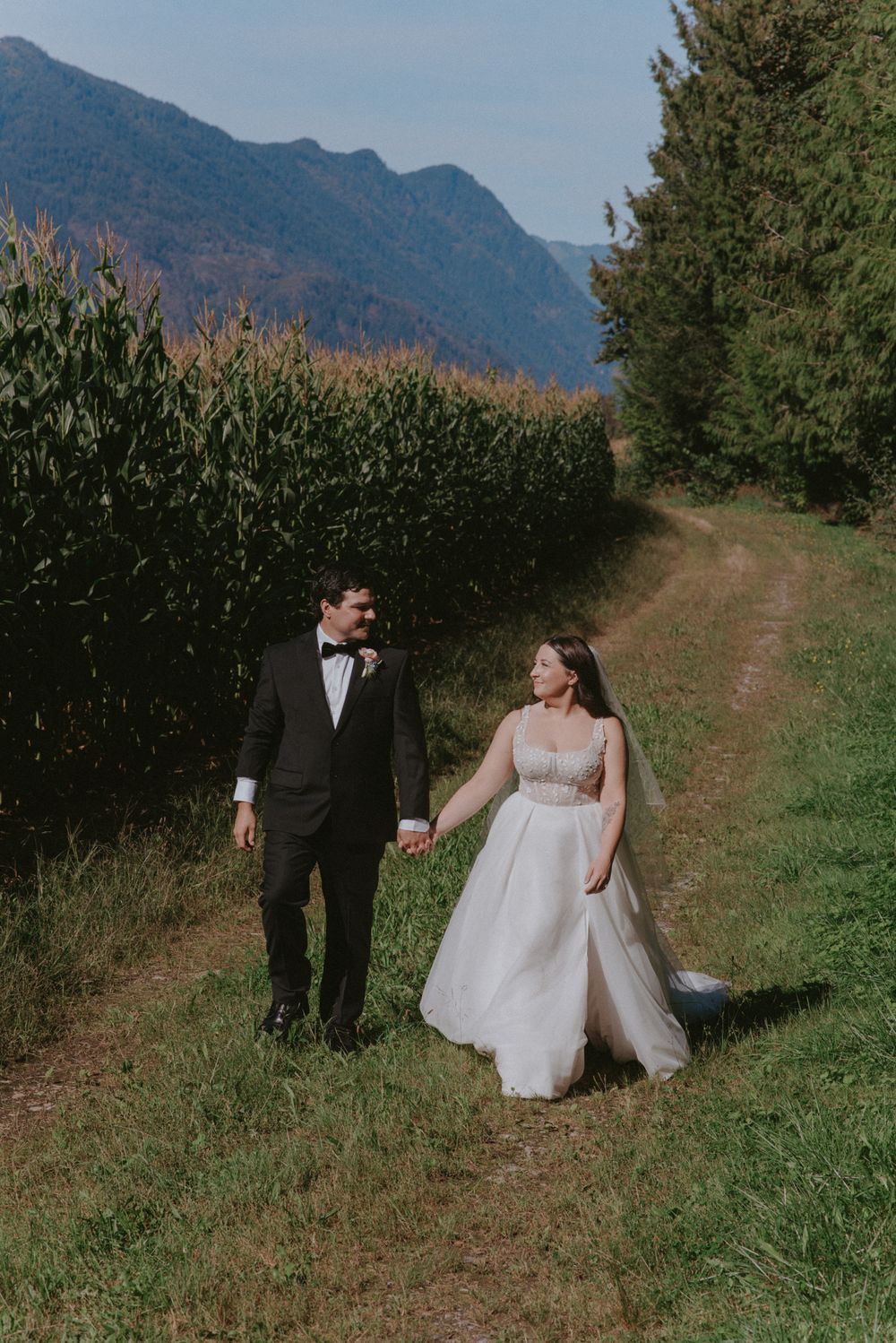 A couple in wedding attire walks along a dirt path with mountains and cornfields in the background on an autumn day.