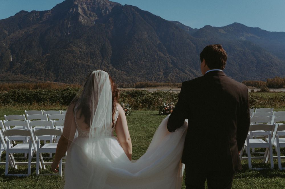A couple in wedding attire stand together on a mountainside with white chairs set up for an outdoor ceremony.