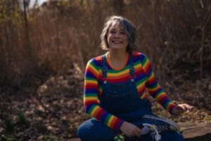Woman in a rainbow shirt and overalls smiling, radiating positive energy and vibrant style.