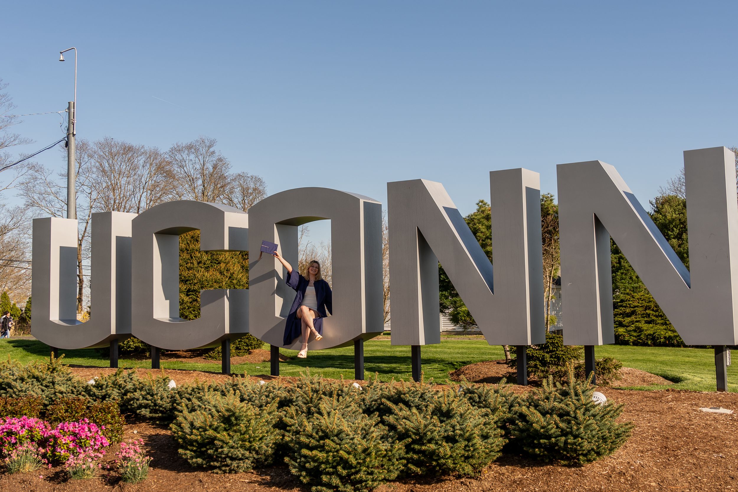 Girl wearing graduation gown raising cap in front of iconic UConn sign on graduation day.