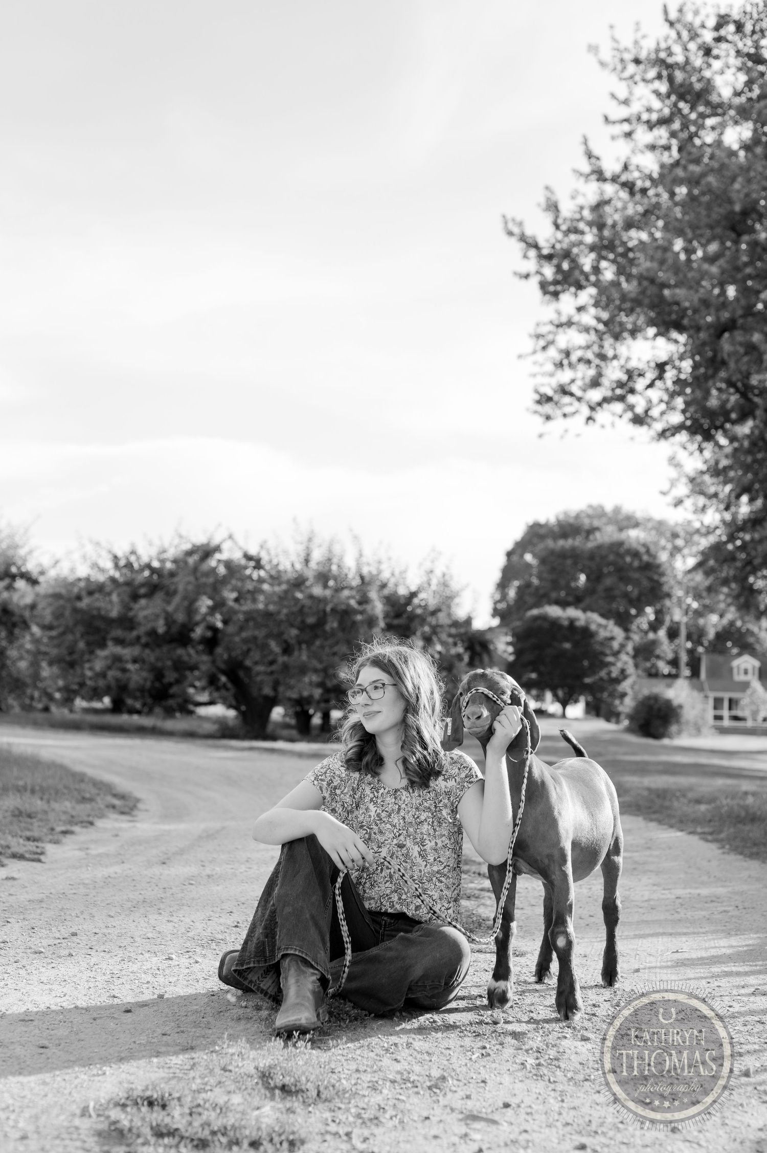 Black and white photo of person sitting with brown goat on rural path.